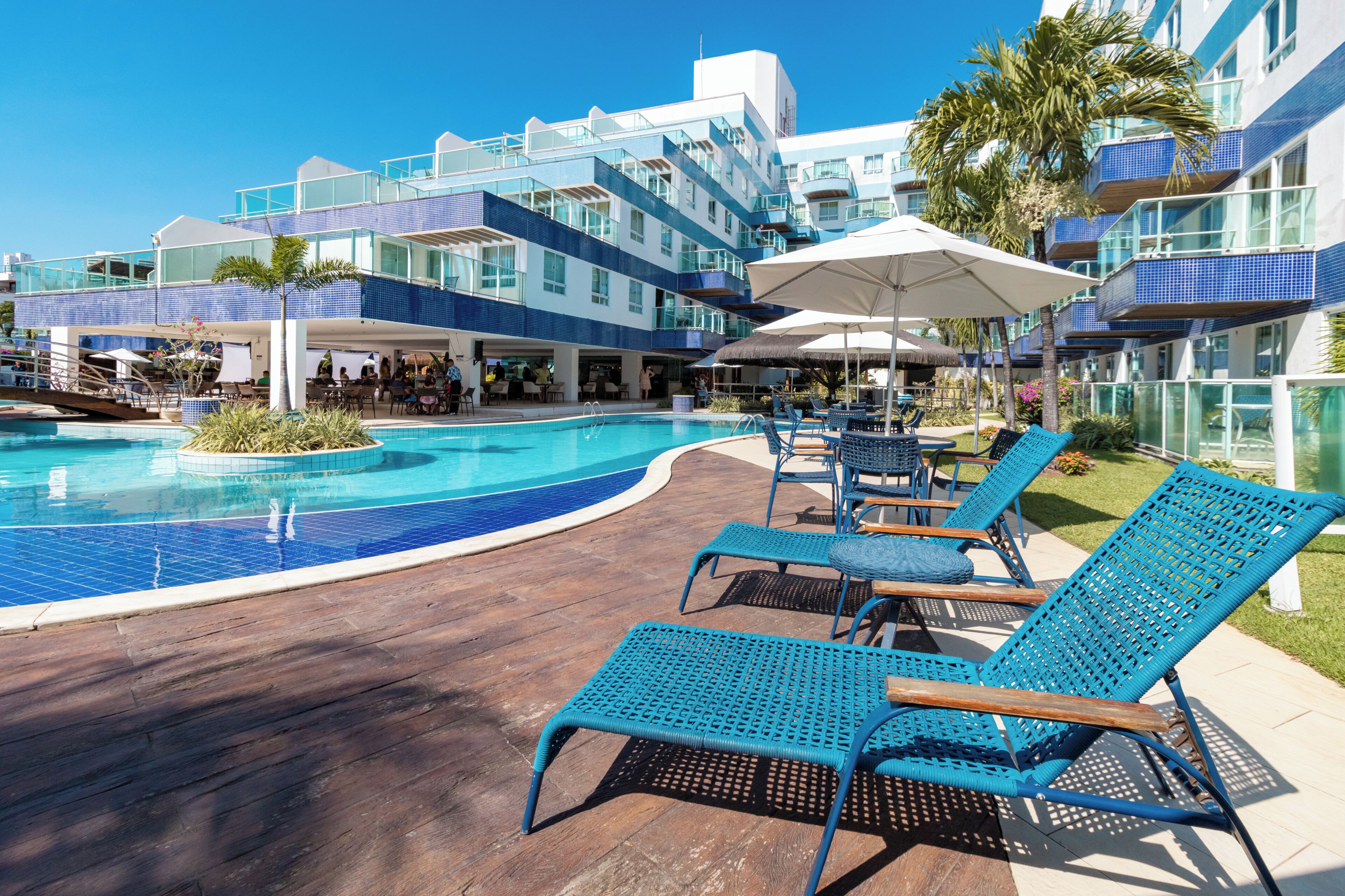 Uma área à beira da piscina com espreguiçadeiras, guarda-sóis e um edifício com várias varandas.