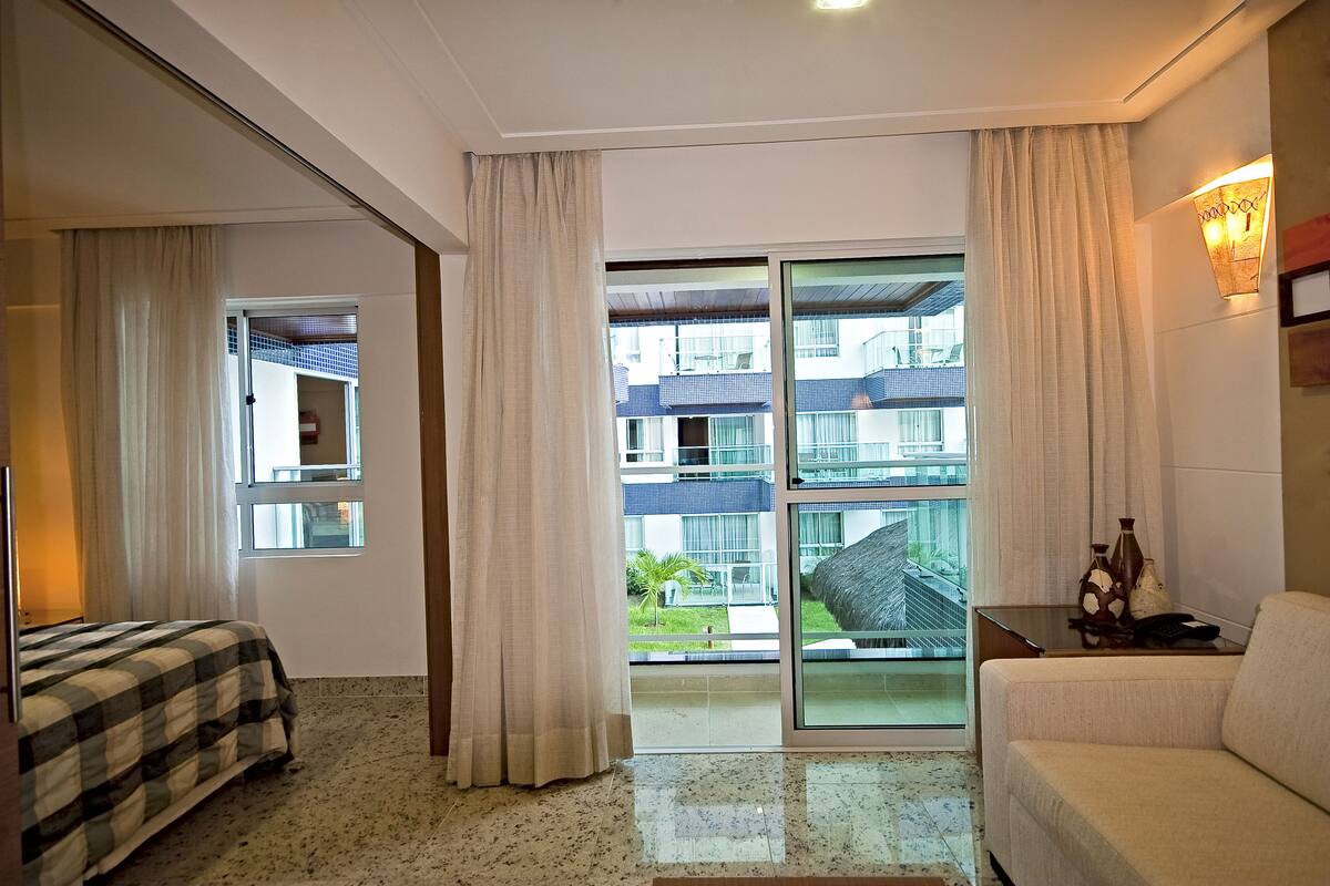 Hotelkamer met een bed, een bank, een tafel met lamp en uitzicht op een gebouw door een glazen deur.