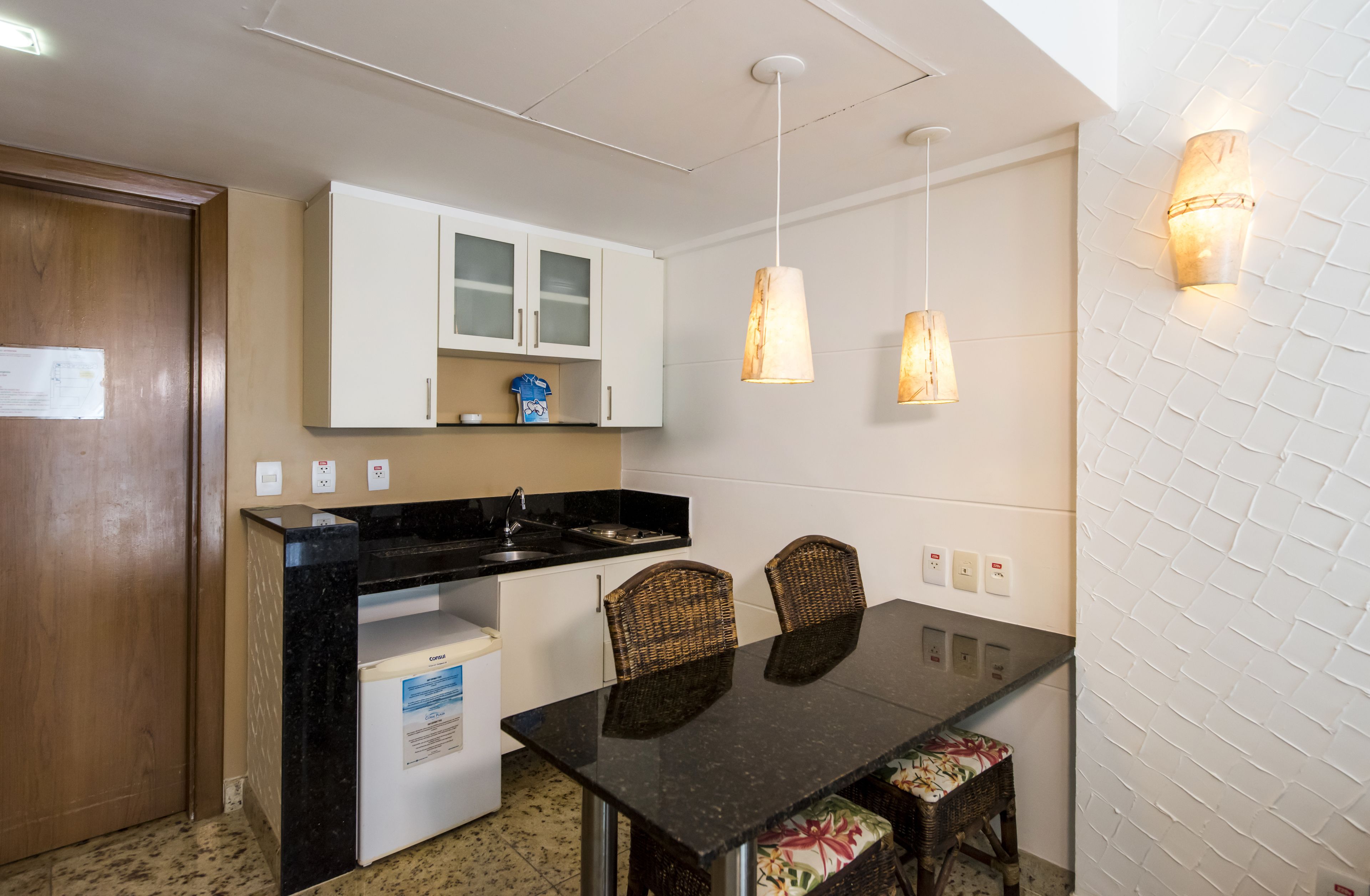 Una cocina compacta con armarios blancos, una encimera negra y una mesa de comedor con sillas de mimbre.