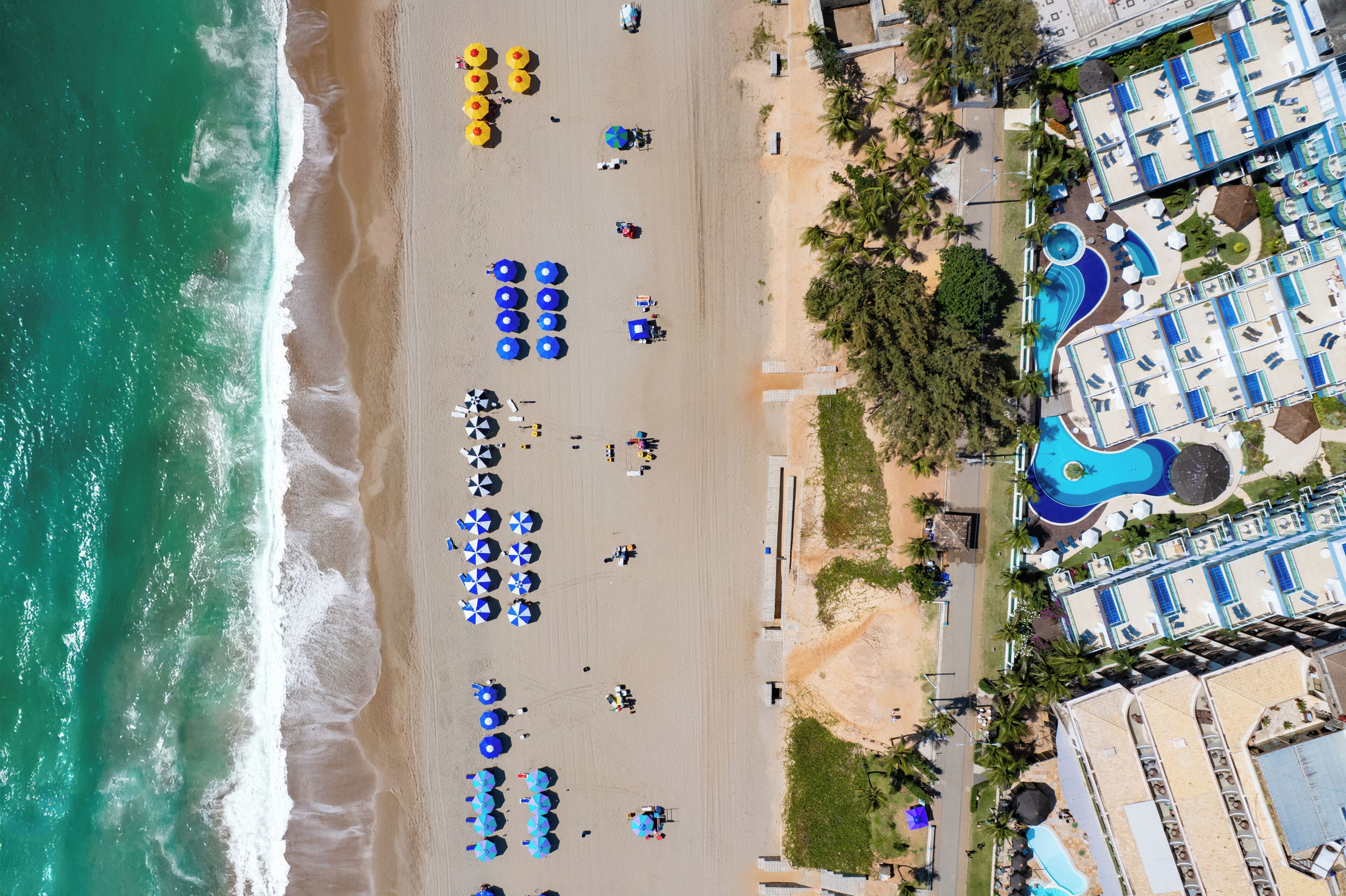 Vista aérea de um resort na praia com várias piscinas, guarda-sóis e uma extensa faixa de areia.