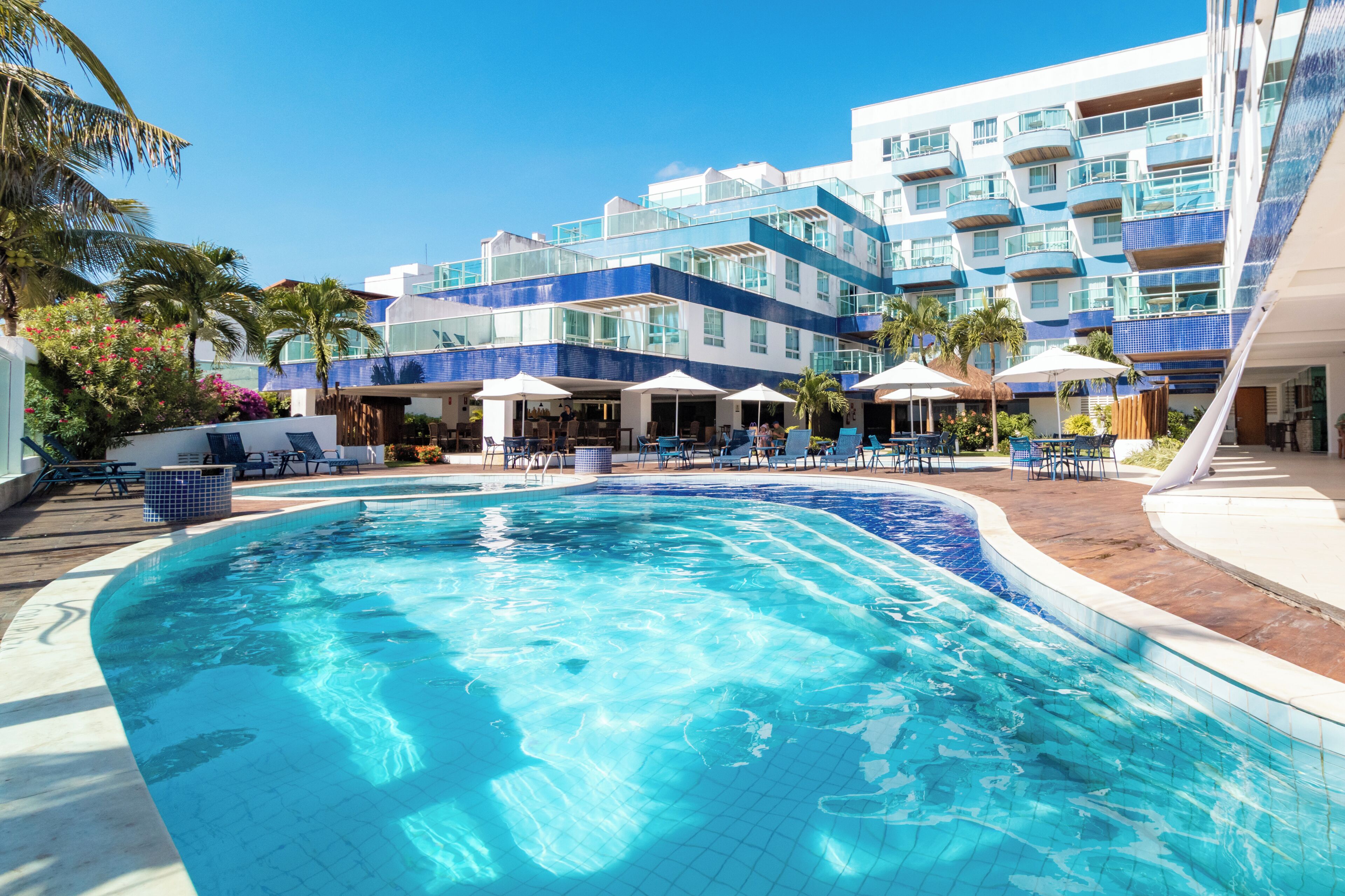 Une piscine avec des chaises longues et des parasols, un immeuble d’hôtel à plusieurs étages doté de balcons, et des palmiers.