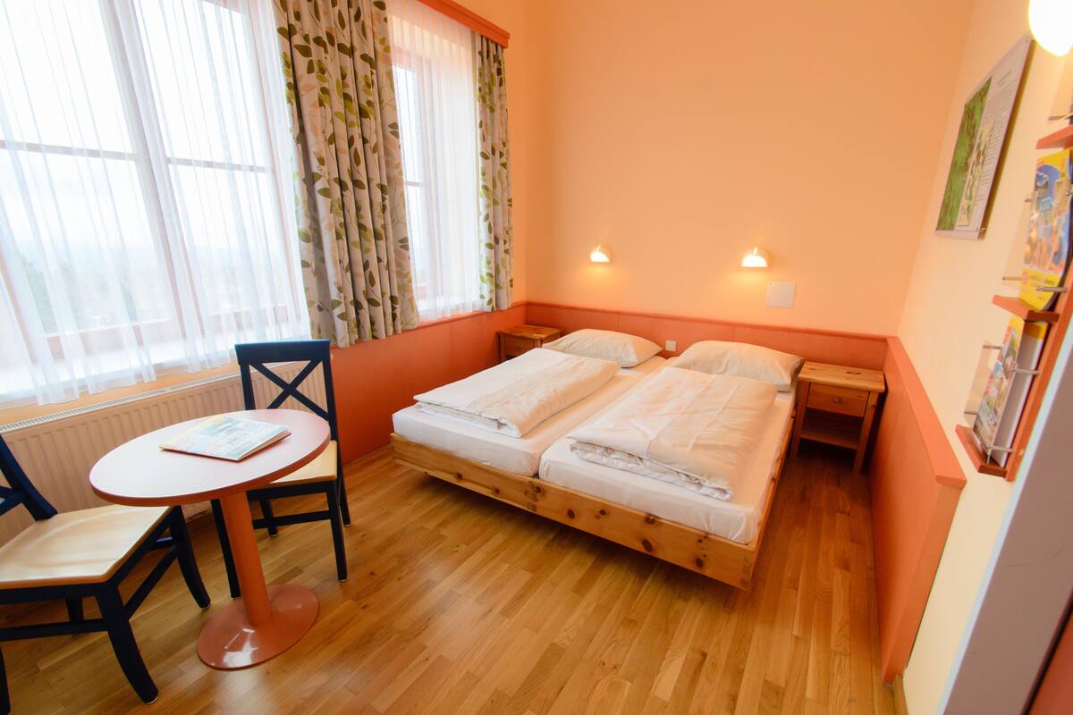 Una habitación con una cama de madera, una mesita redonda con una silla, una ventana con cortinas ligeras y una lámpara fijada a la pared.