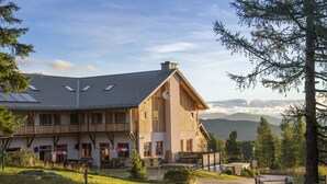 A large wooden building with multiple balconies and a sloped roof, surrounded by a grassy area and trees. -  JUFA Hotel Hochrindl (Albeck)