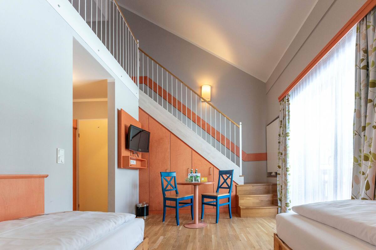 Habitación de hotel con escalera, una cama, dos sillas, una mesita con botellas y un televisor en la pared.