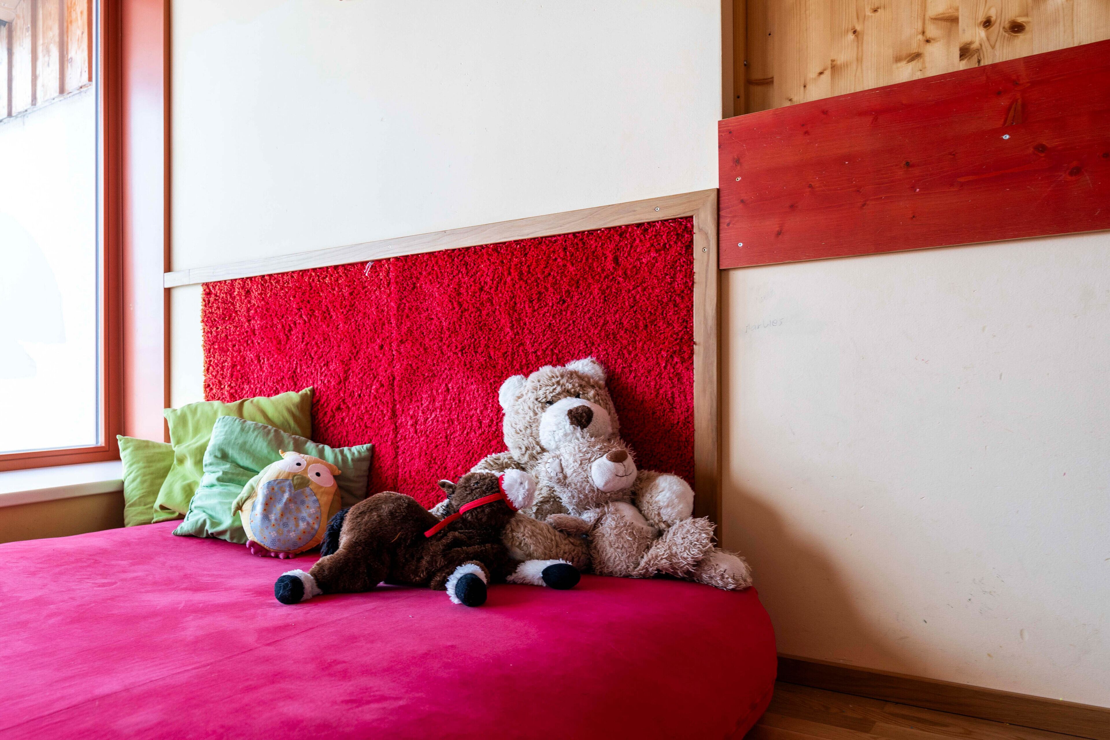 Un lit avec une tête de lit rouge et des peluches, dont un ours en peluche et une chouette, posées sur une couverture rose.