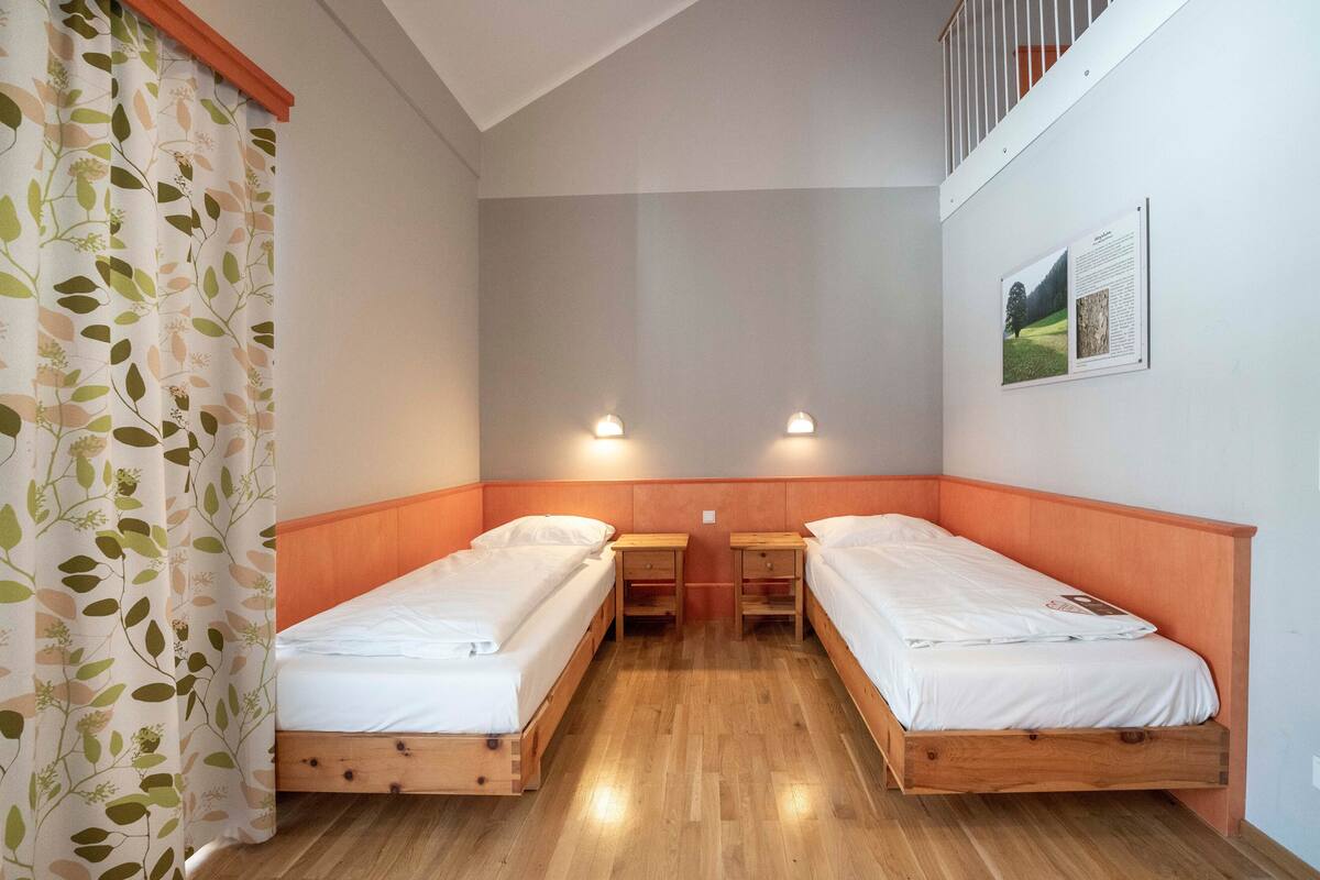 Una habitación con dos camas de madera, una mesita de noche de madera, una cortina blanca con estampado de hojas y una lámpara fijada a la pared.