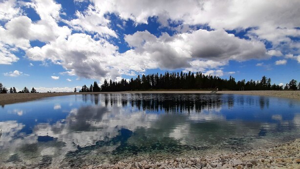 A calm lake with clear reflections of the sky and trees, surrounded by a rocky shoreline and a forest in the background. -  JUFA Hotel Hochrindl (Albeck)