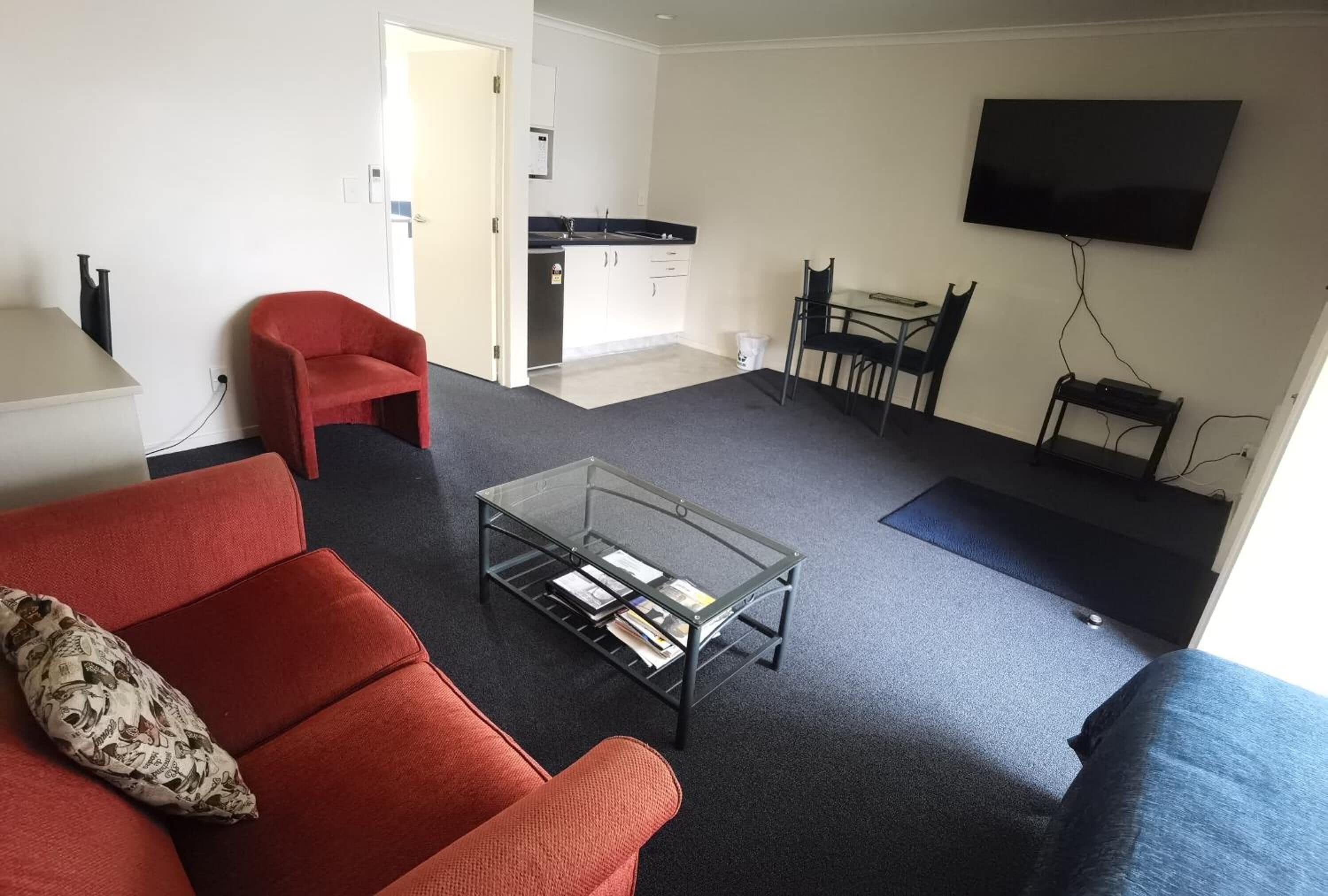 A living room with a red sofa, a glass coffee table, a TV, and a kitchen area in the background.