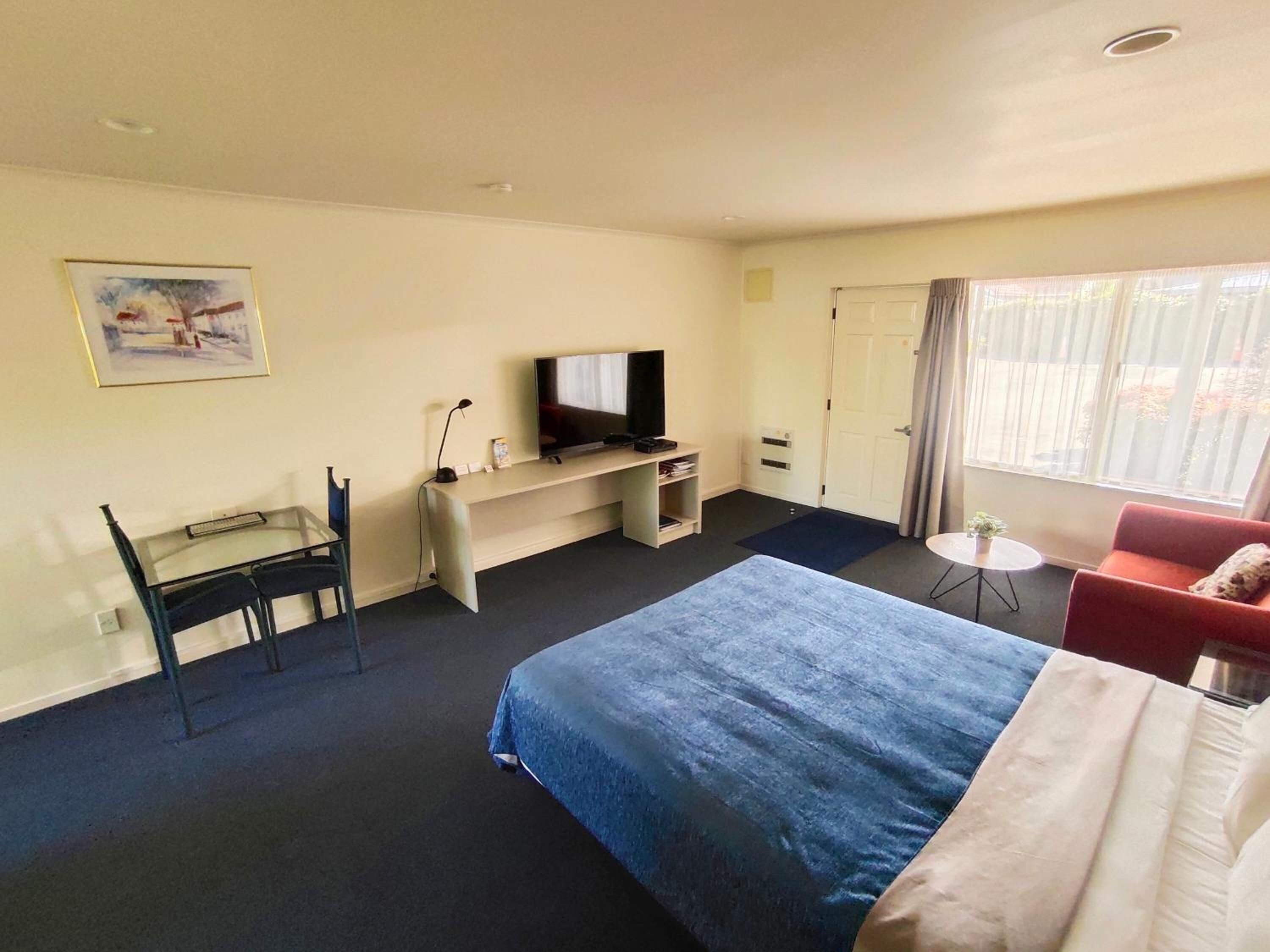 A hotel room with a bed, a desk with a television, a chair, a small table, and a window with curtains.