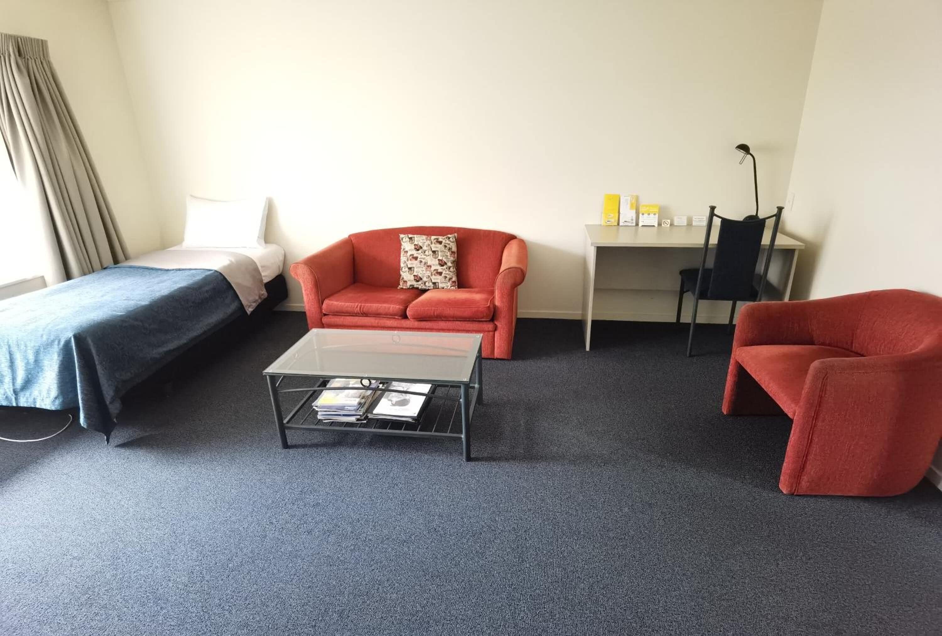 A hotel room with a bed, two red armchairs, a small table with magazines, a desk with a lamp, and a window with curtains.
