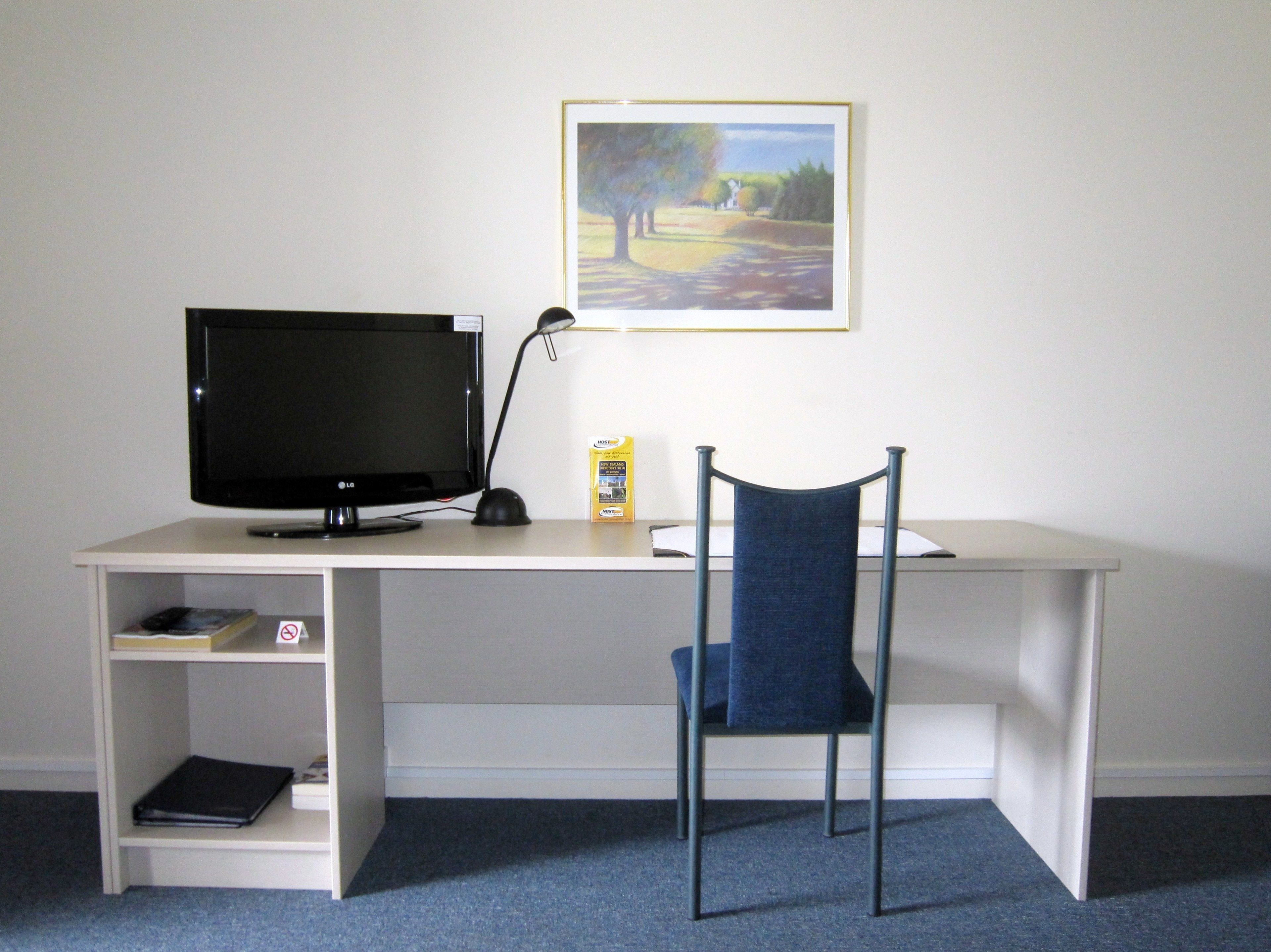 A white desk with a black monitor, a lamp, a chair, and a framed landscape painting on the wall.