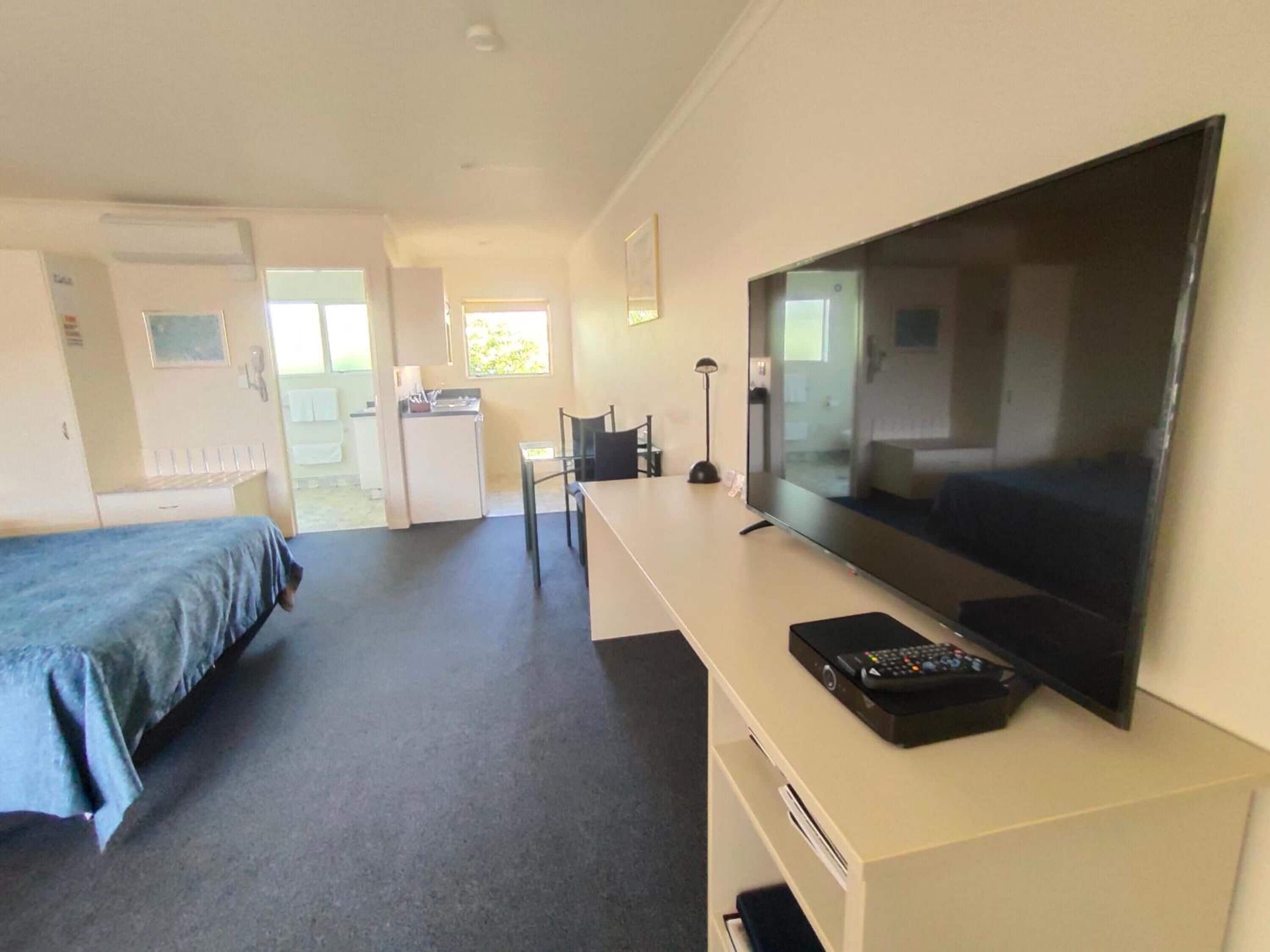 A hotel room with a bed, a desk with a TV, a chair, a desk lamp, and a small kitchenette.