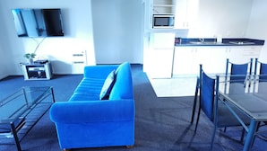 Een moderne woonkamer met een blauwe bank, een glazen salontafel en een flatscreen televisie aan de muur.