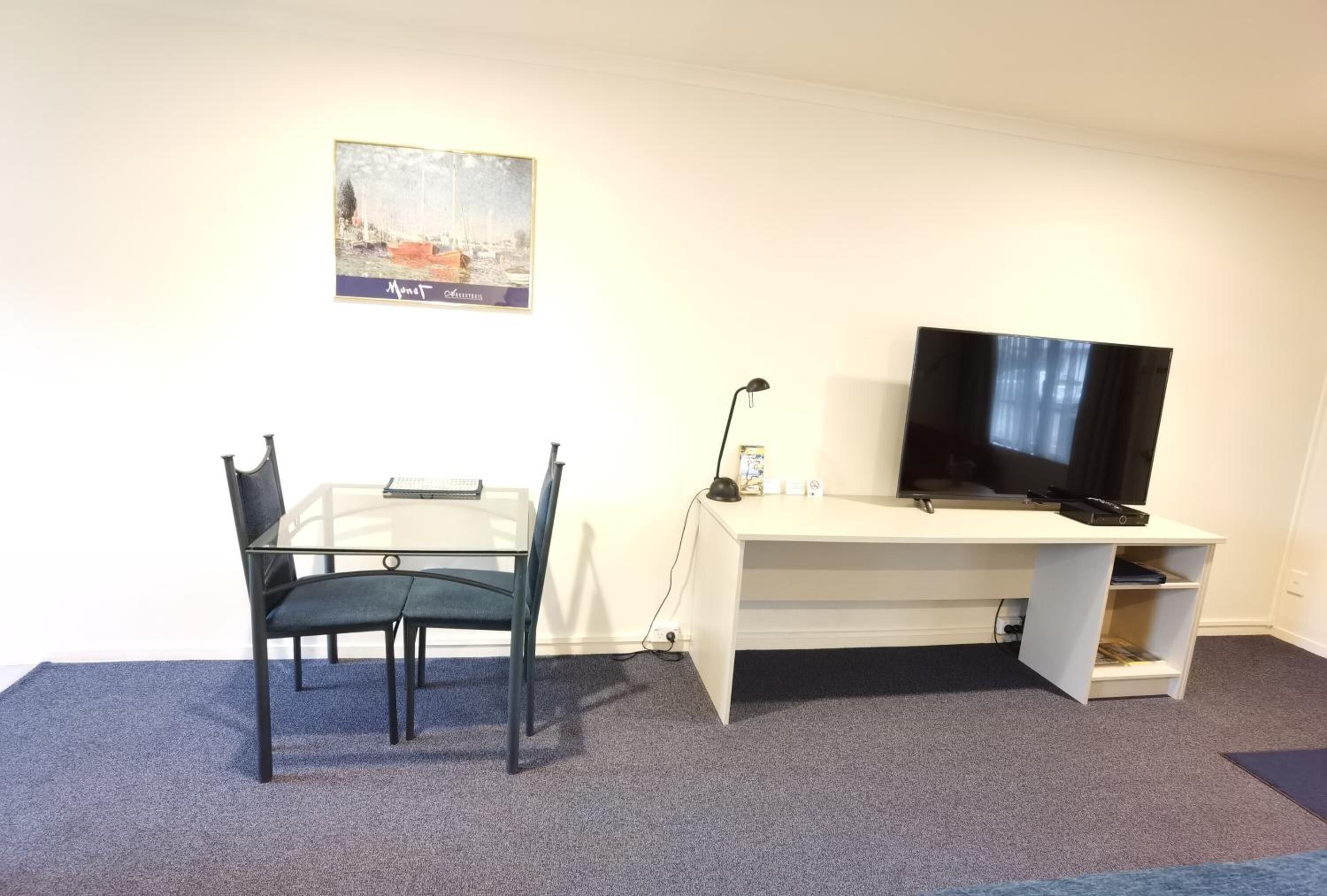 A room with a glass table and chairs, a white desk with a television, and a painting on the wall.