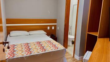 Une chambre d’hôtel avec un lit, une table de chevet, un téléphone, une armoire en bois et une salle de bain visible à travers une porte ouverte.