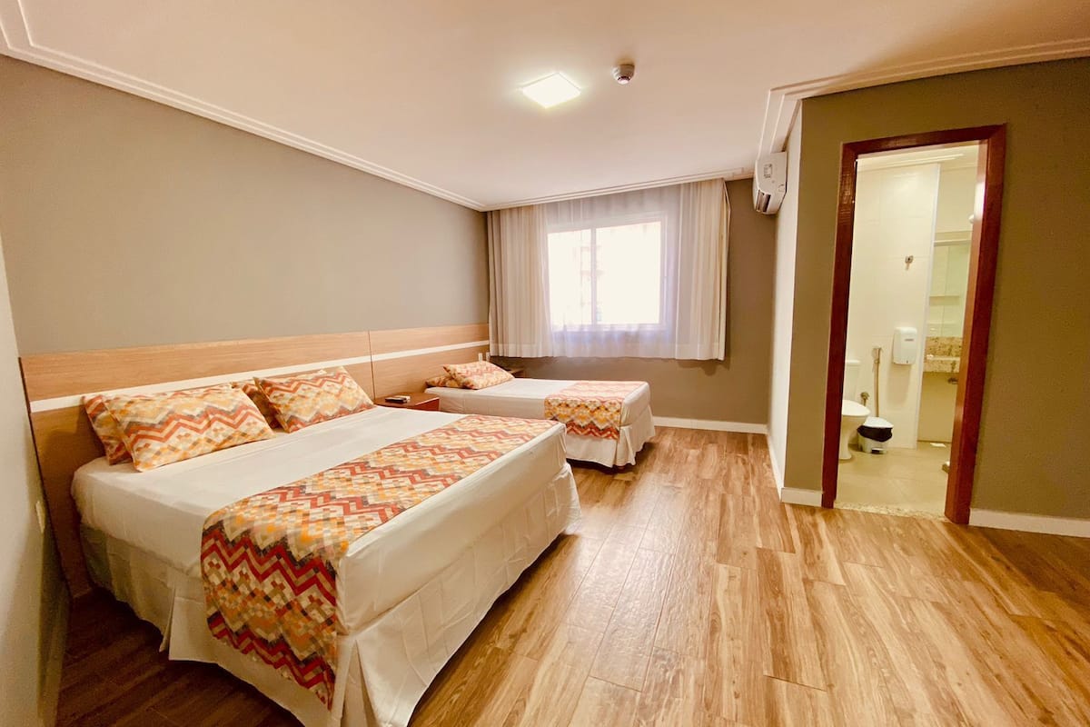 A hotel room with two beds, a wooden floor, a window with curtains, and a bathroom visible through an open door.