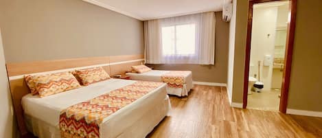 A hotel room with two beds, a wooden floor, a window with curtains, and a bathroom visible through an open door.