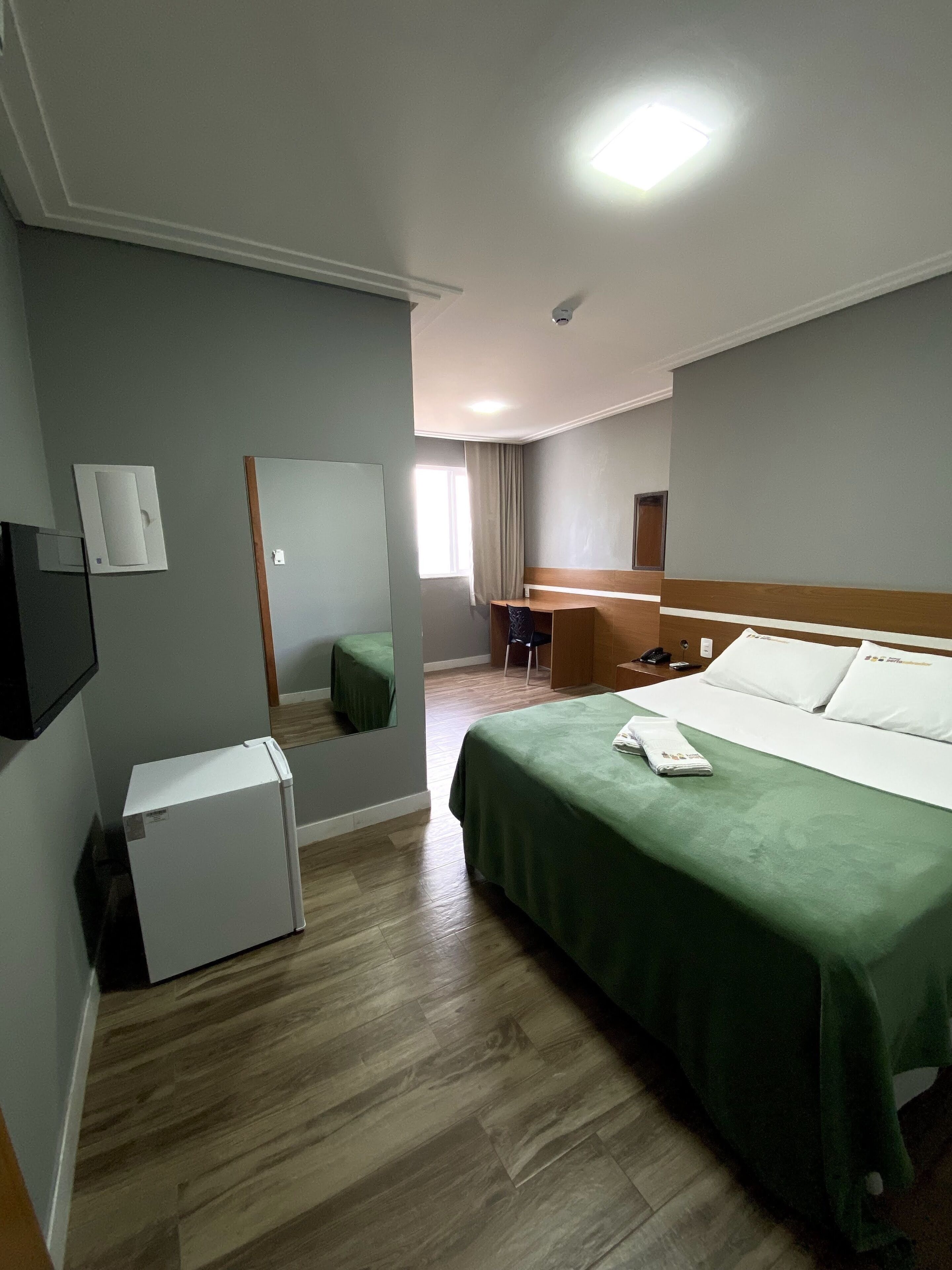 A hotel room with a bed, a small refrigerator, a desk, and a mirror.