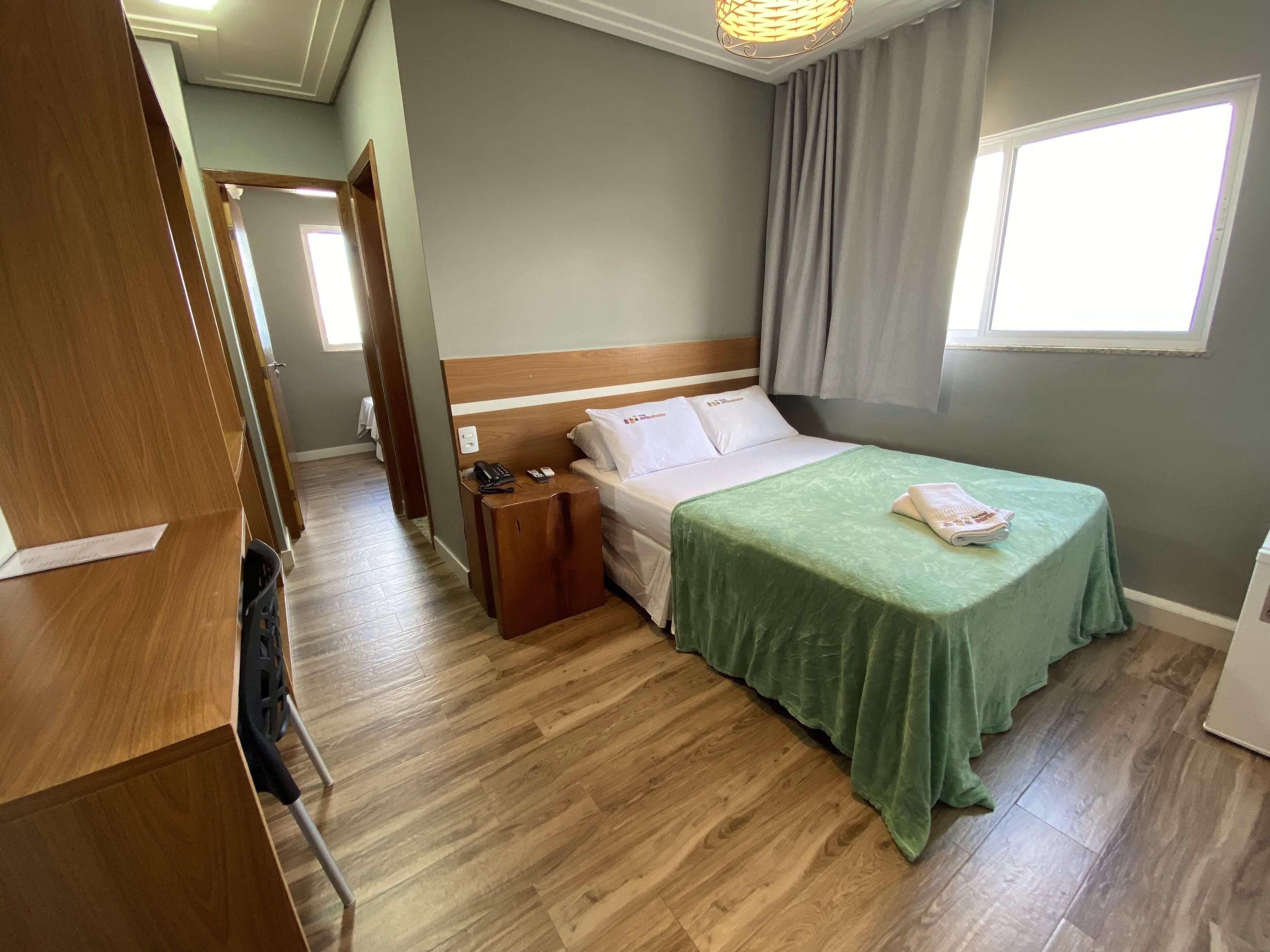 A hotel room with a wooden floor, a bed with a green bedspread, a bedside table with a phone, and a window with curtains.