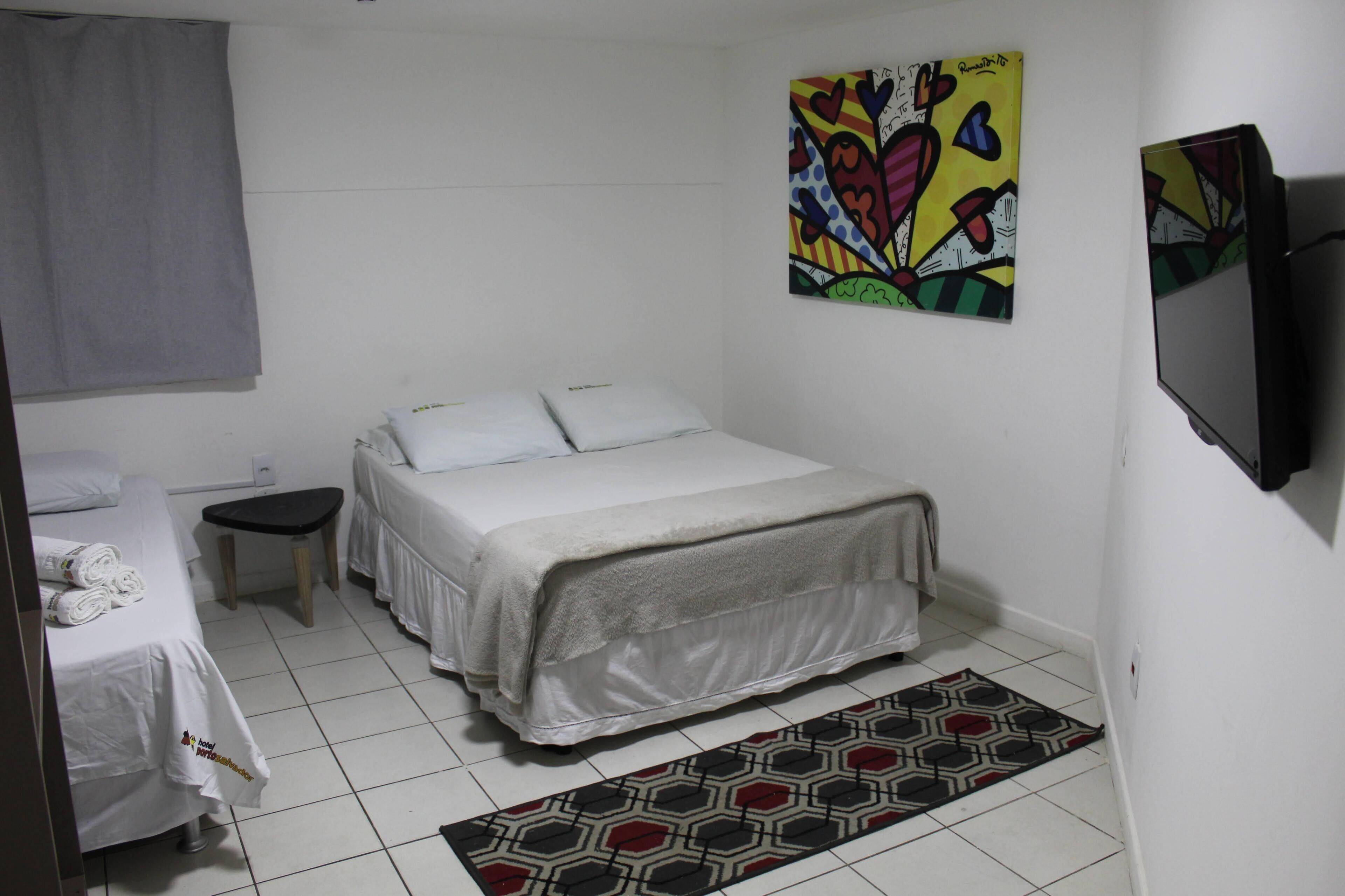Quarto de hotel com cama, uma mesinha, uma cadeira, televisão e uma obra de arte colorida na parede.