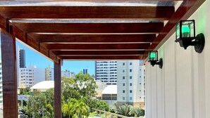 A balcony with a wooden floor and white walls, overlooking a cityscape with buildings and palm trees.