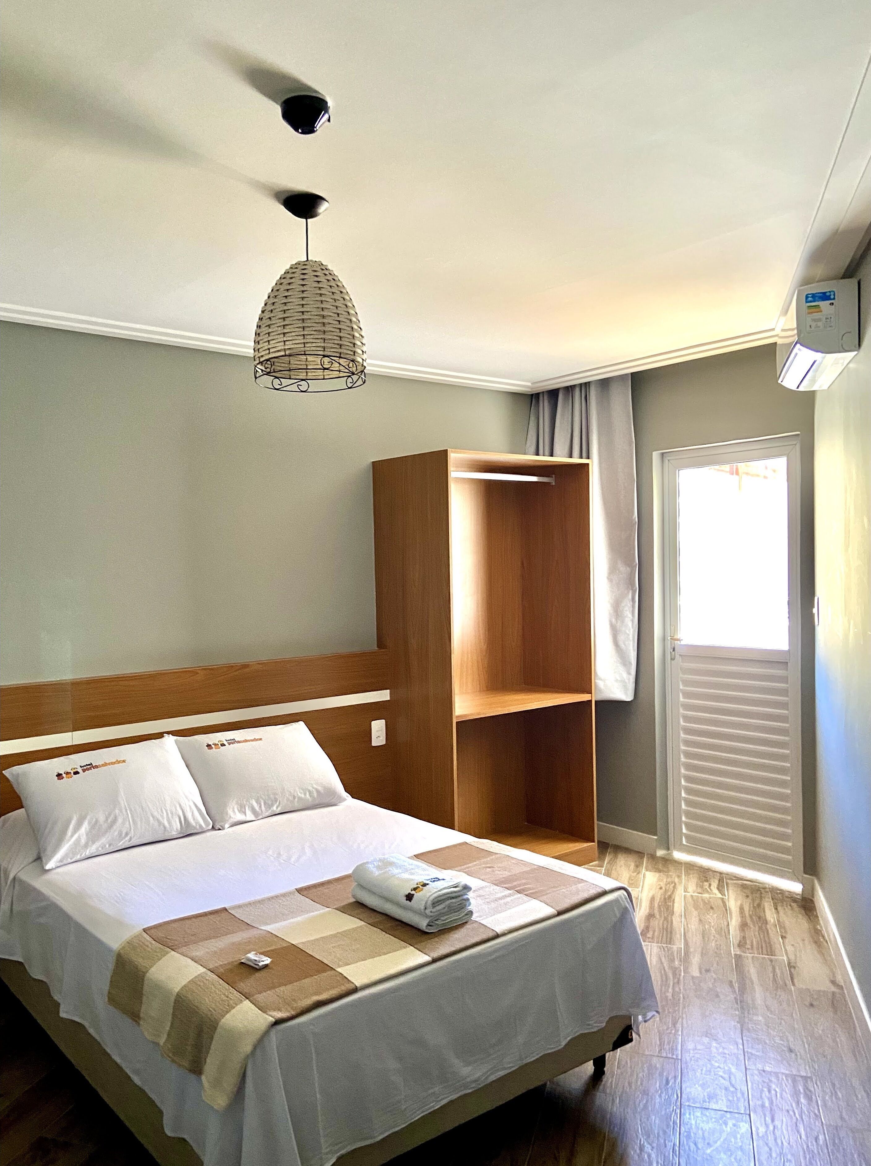 Una habitación de hotel con una cama, un armario de madera, una lámpara colgante y una ventana con cortinas.