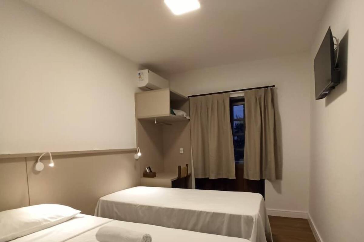 Een hotelkamer met twee bedden, een plafondlamp, een aan de muur bevestigde televisie en een raam met gordijnen.