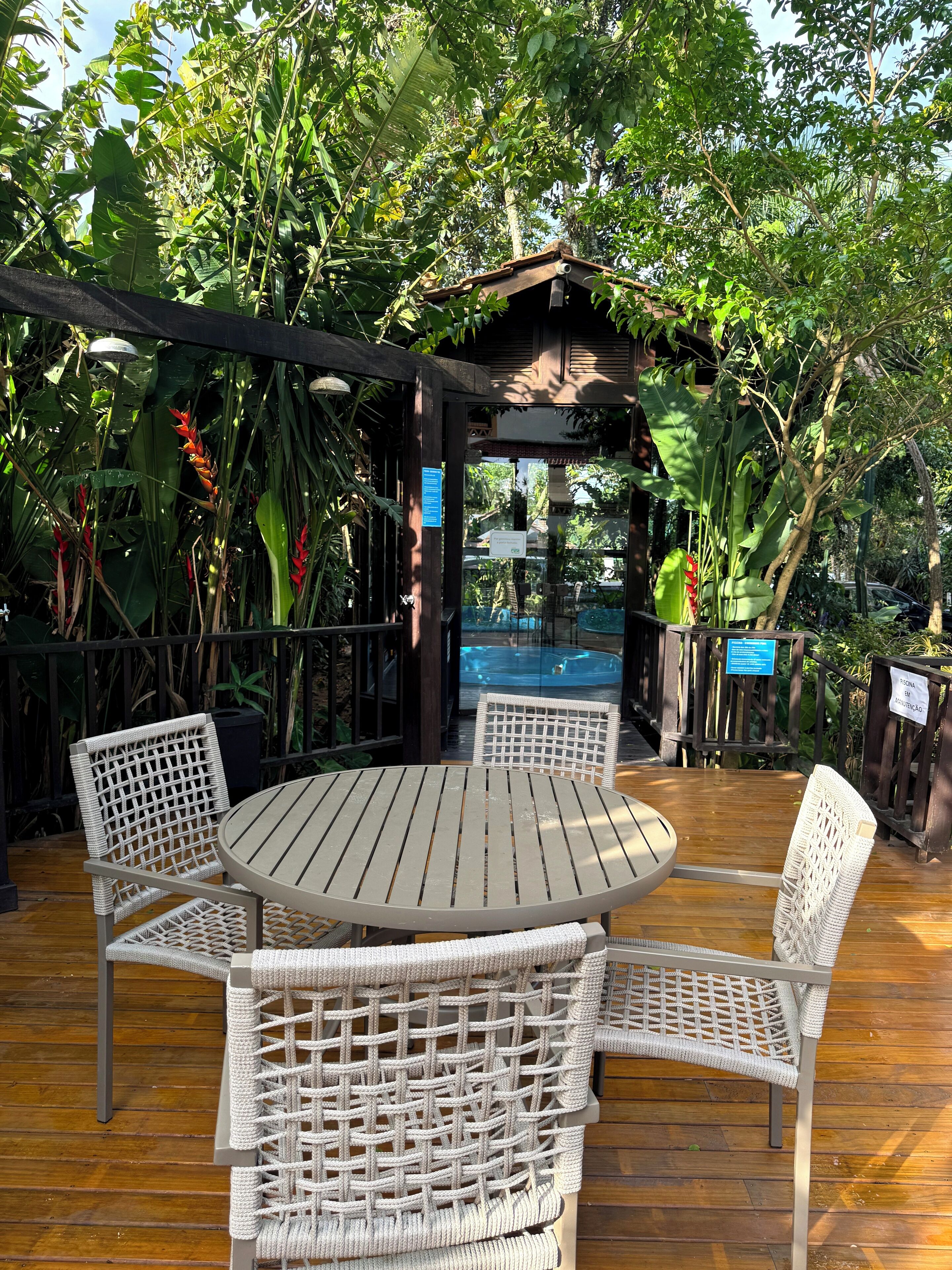 A round wooden table with four wicker chairs, surrounded by lush greenery and a wooden deck.