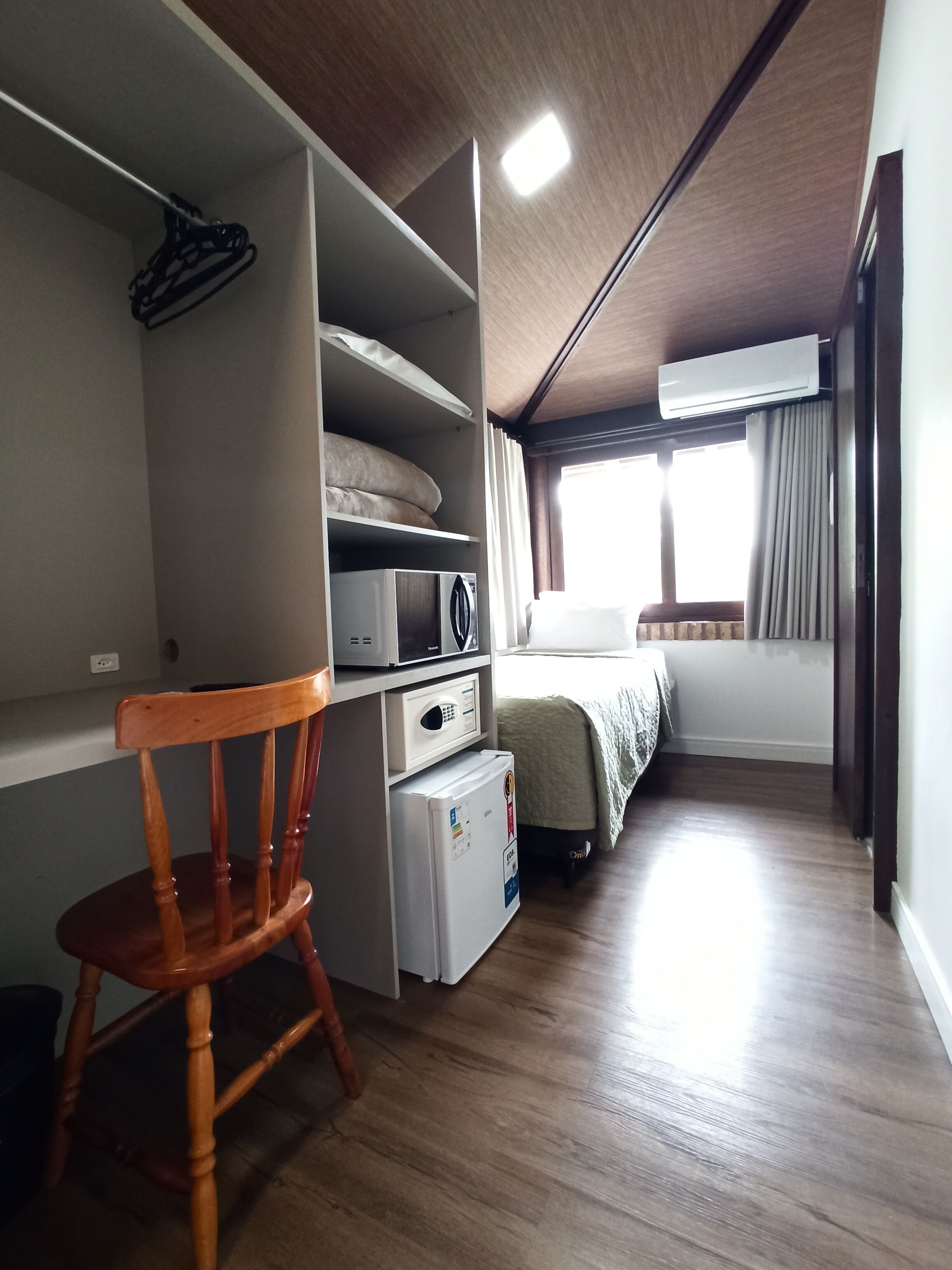 Una habitación pequeña con suelo de madera, una cama con cubrecama blanco, un refrigerador blanco pequeño, un microondas y una silla de madera.