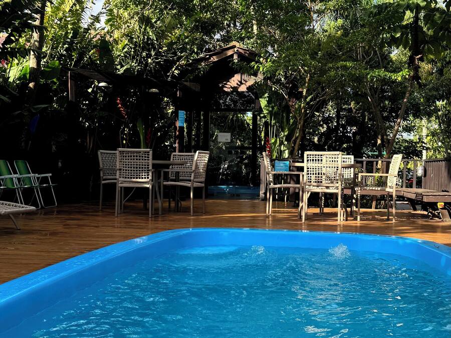 Una piscina rodeada de sillas y mesas de madera, con abundante vegetación por encima.