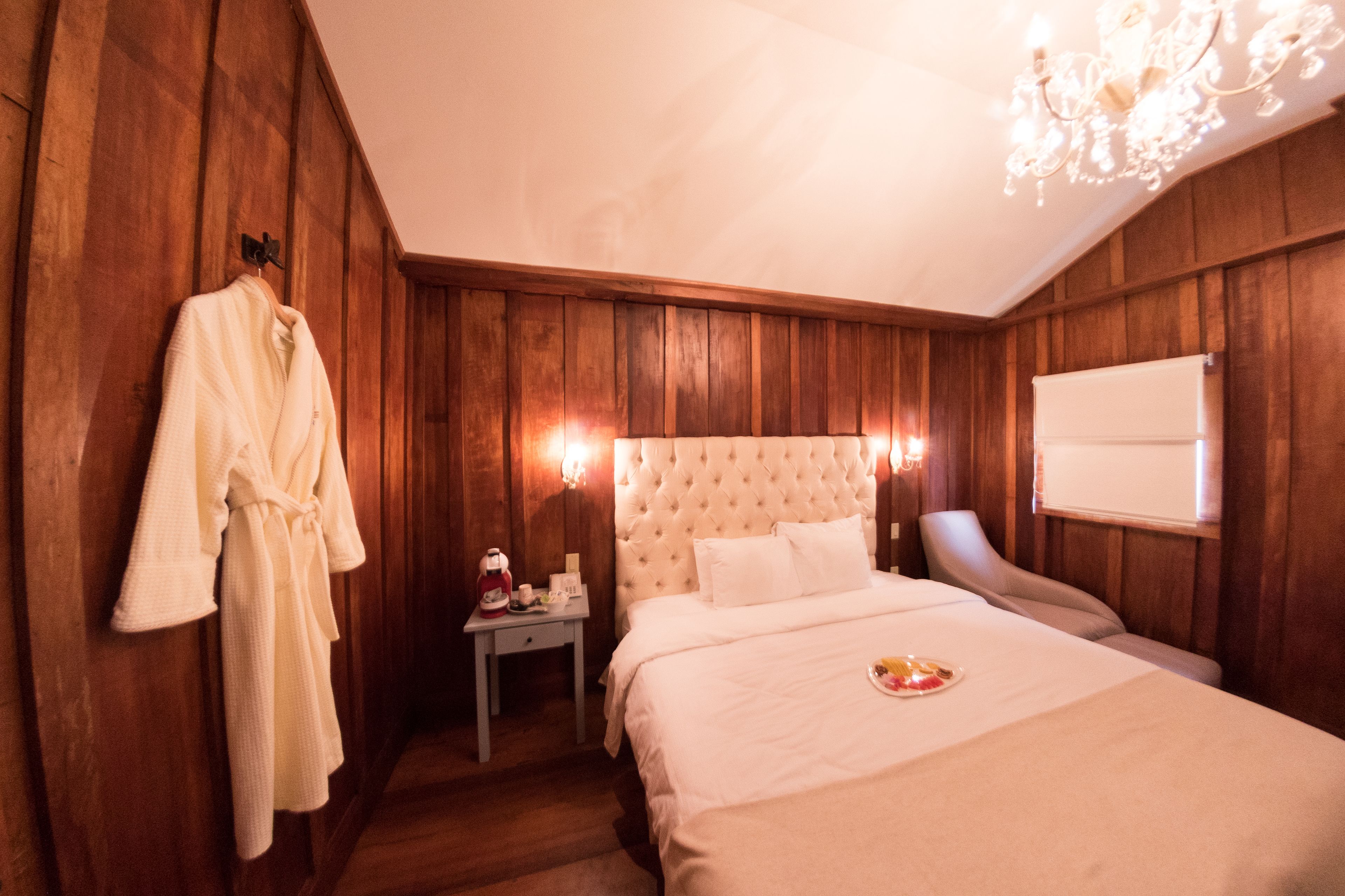 A hotel room with wooden paneling, a large bed with white linens, a nightstand with a lamp, and a robe hanging on the wall.