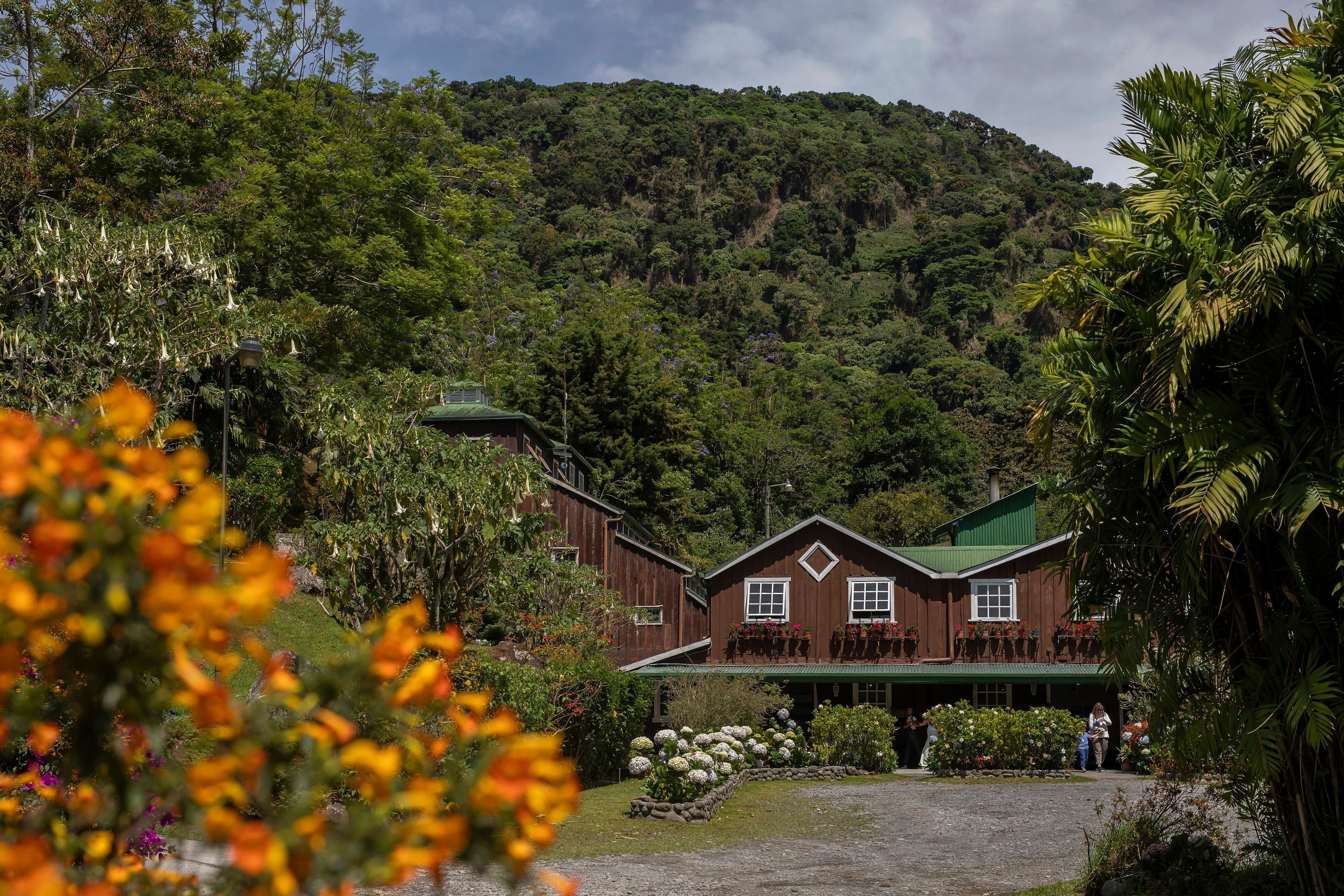 Un edificio rústico de madera con techo verde, rodeado de exuberante vegetación y montañas al fondo.