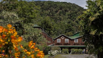 Un edificio rústico de madera con techo verde, rodeado de exuberante vegetación y montañas al fondo.