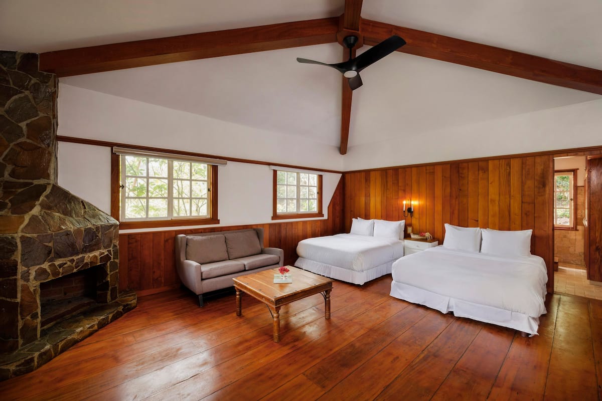 A room with a stone fireplace, wooden ceiling beams, a ceiling fan, a sofa, two beds, and large windows.