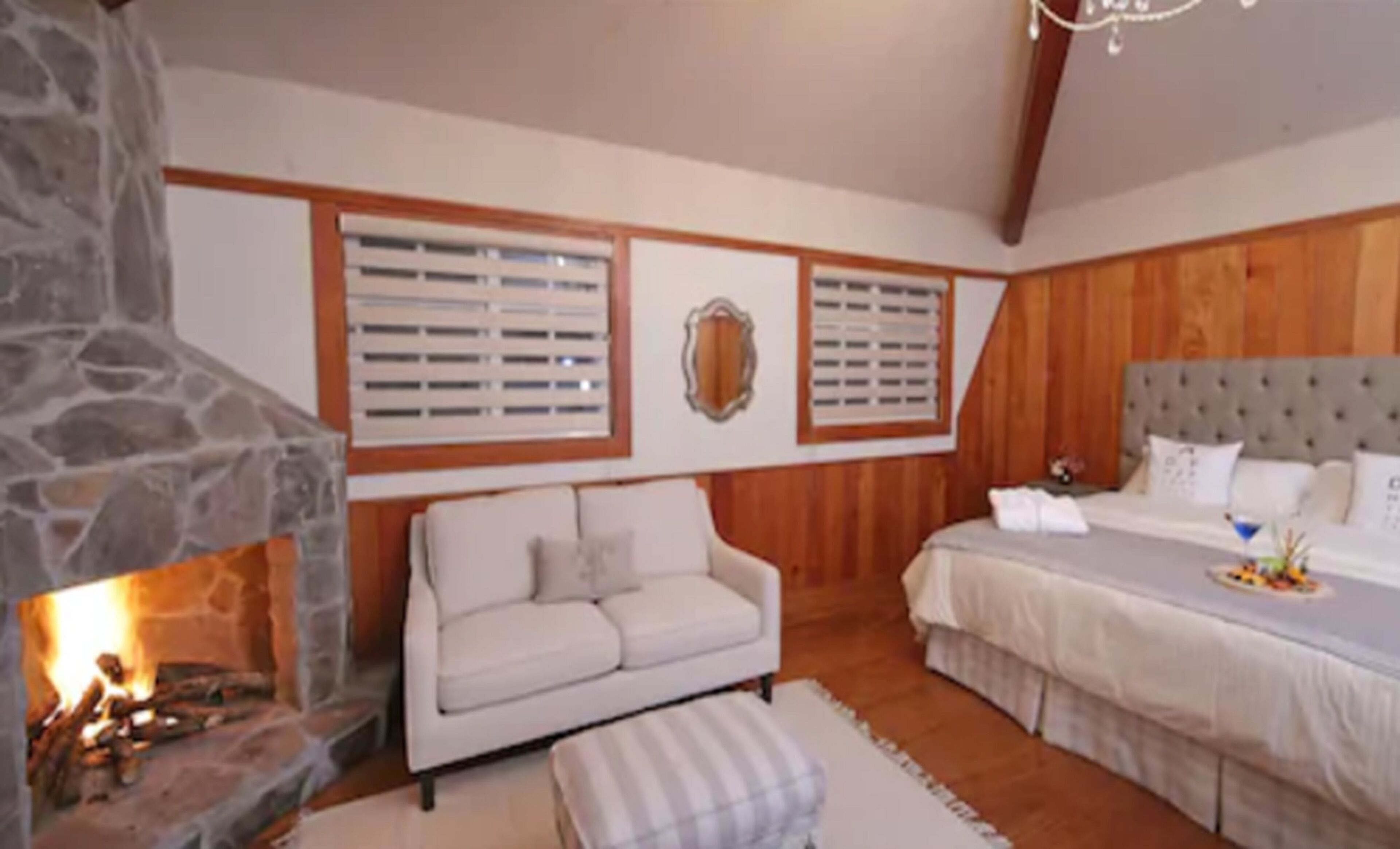 Un dormitorio acogedor con chimenea de piedra, un sofá blanco, cama con cabecero gris y paneles de madera en las paredes.