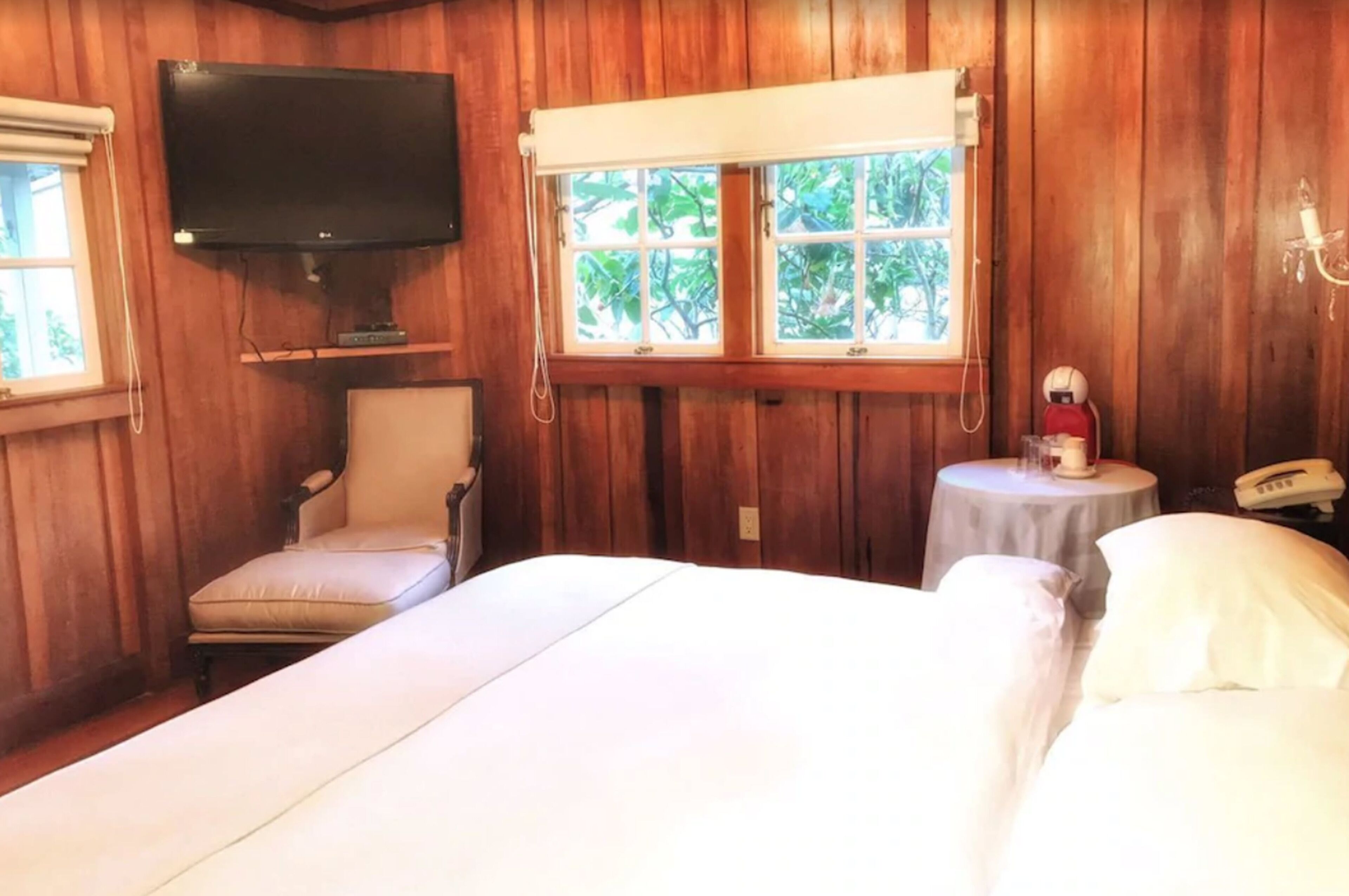 Habitación con paneles de madera, una cama, una silla, un televisor, una ventana con vistas a árboles y una mesita de noche con una lámpara y un teléfono.