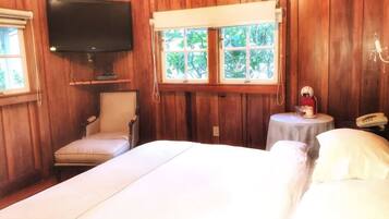 Habitación con paneles de madera, una cama, una silla, un televisor, una ventana con vistas a árboles y una mesita de noche con una lámpara y un teléfono.