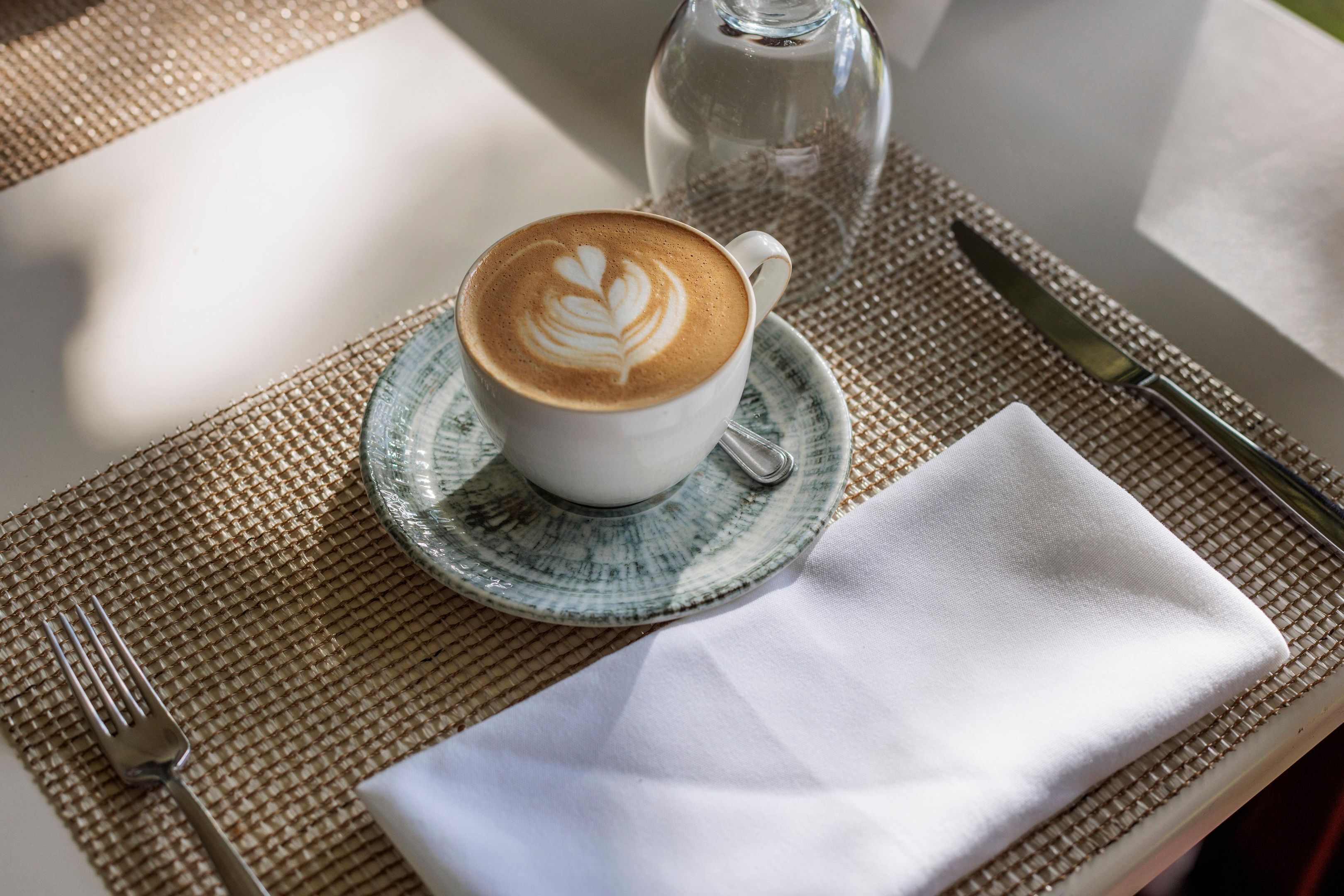 Una taza de café con arte de latte sobre un platito, sobre una mesa con un mantel individual, una botella de vidrio y cubiertos.