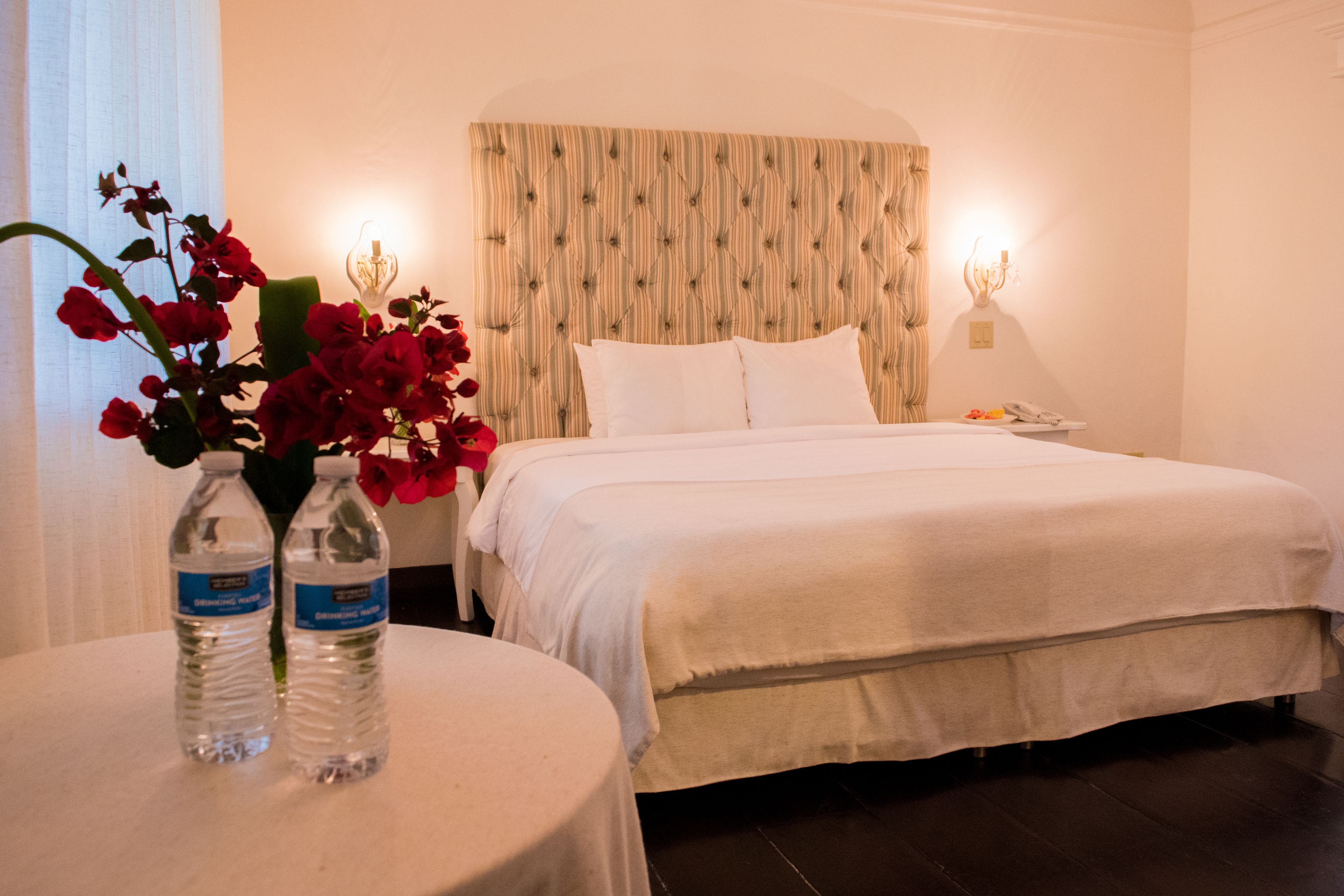 Una cama bien hecha con un cabecero a rayas, una mesita de noche con dos botellas de agua, y un jarrón con flores rojas.