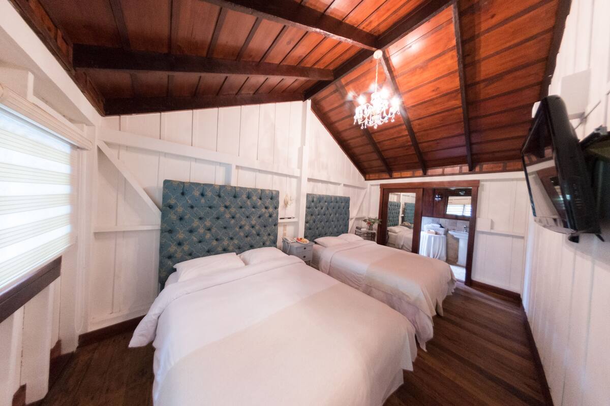 A room with two beds, wooden ceiling beams, a chandelier, and a television on the wall.