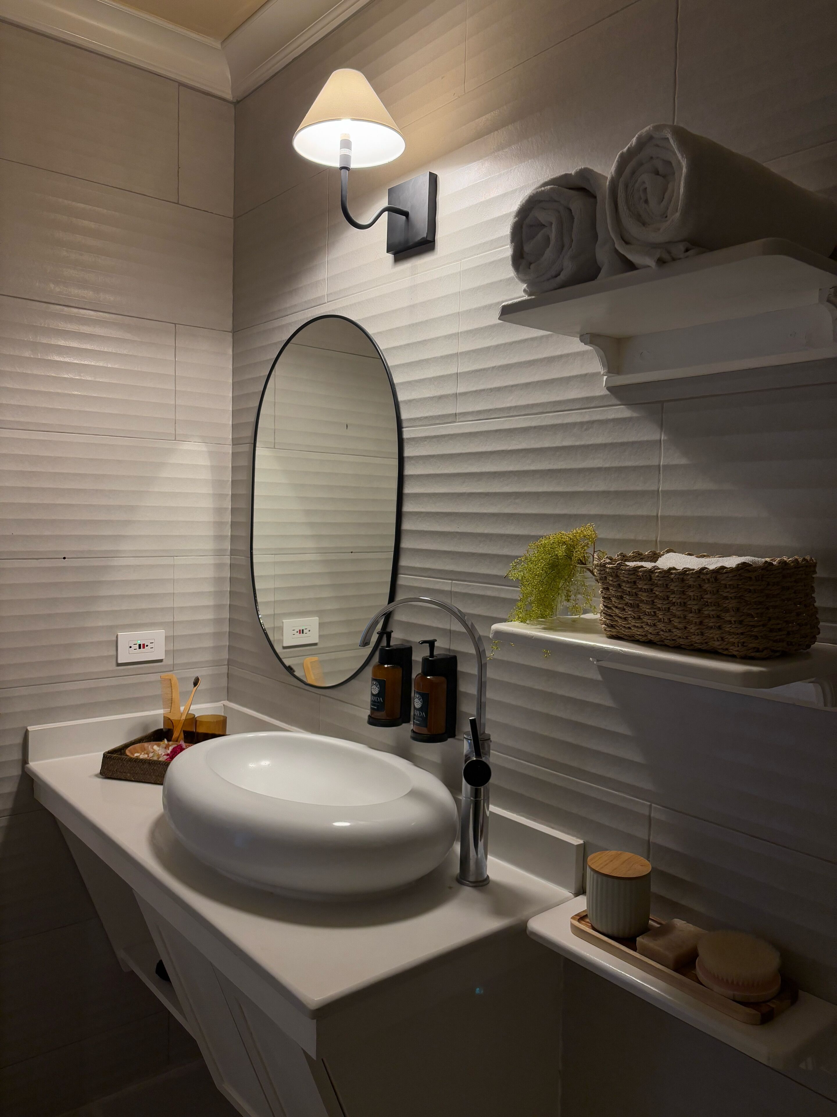 A modern bathroom with a large oval mirror, a white sink, and a wall-mounted light fixture.