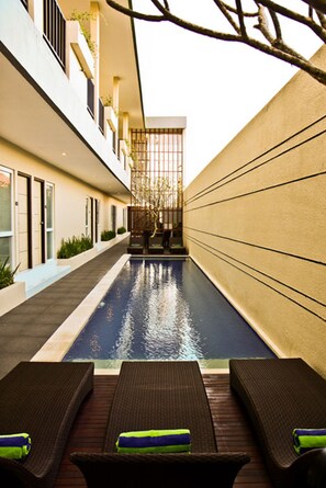 A narrow indoor swimming pool with lounge chairs and a glass-enclosed area with plants. - Eazy Suite (Legian)