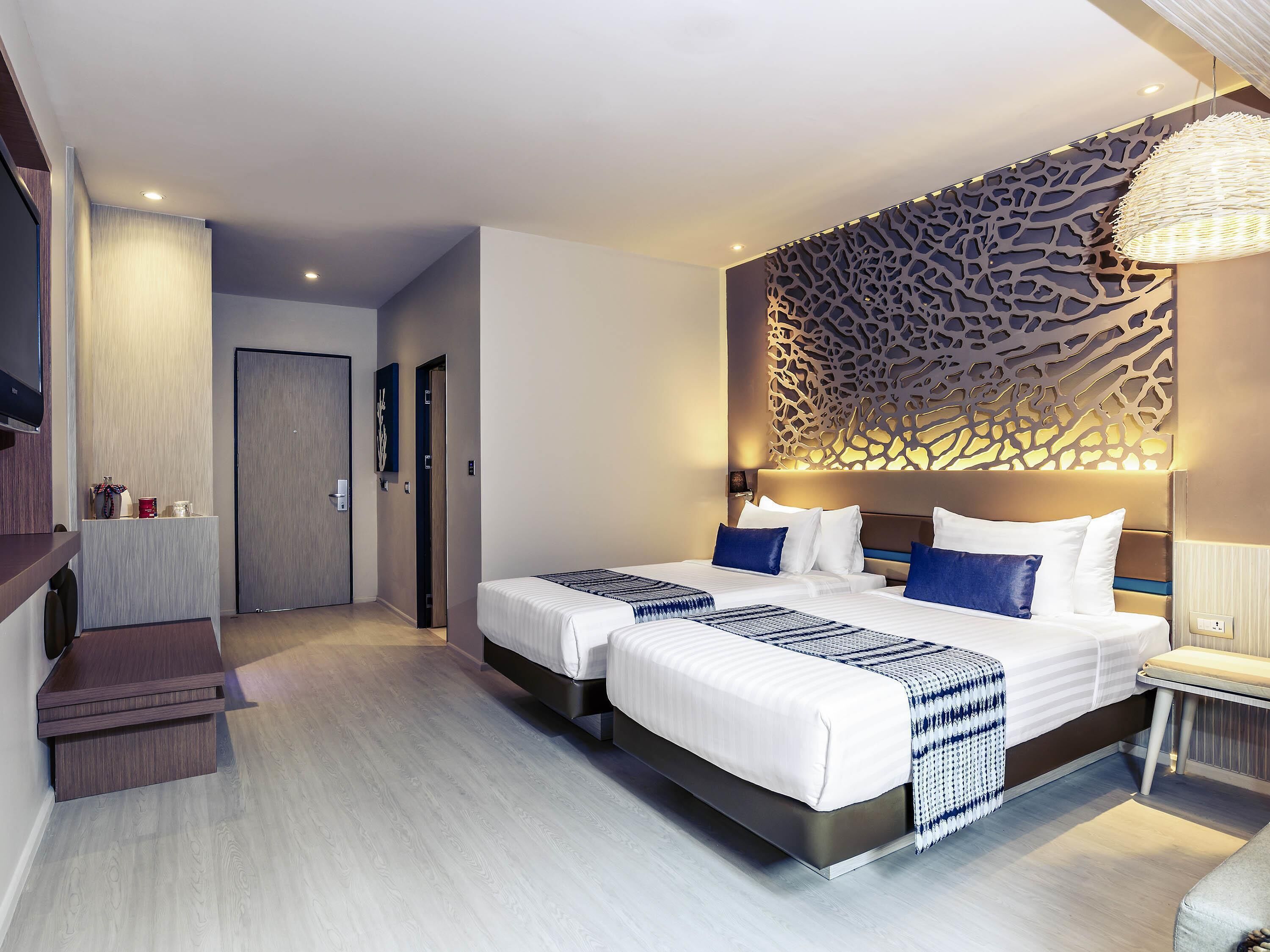 A modern hotel room with two beds, a decorative wall panel, a bedside table with a lamp, and a TV mounted on the wall.