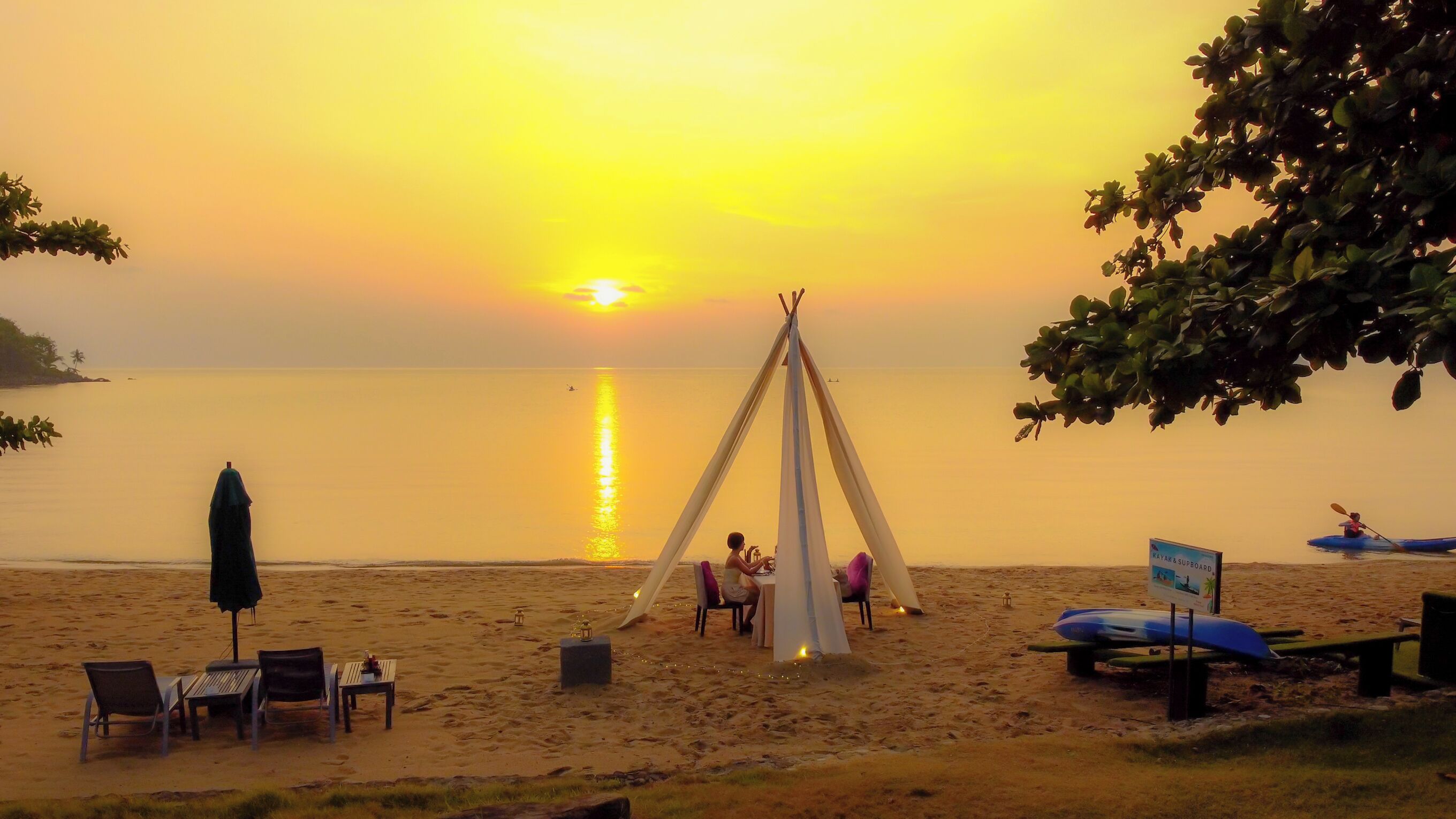 A beach scene with a teepee, chairs, a table, kayaks, and a sign.