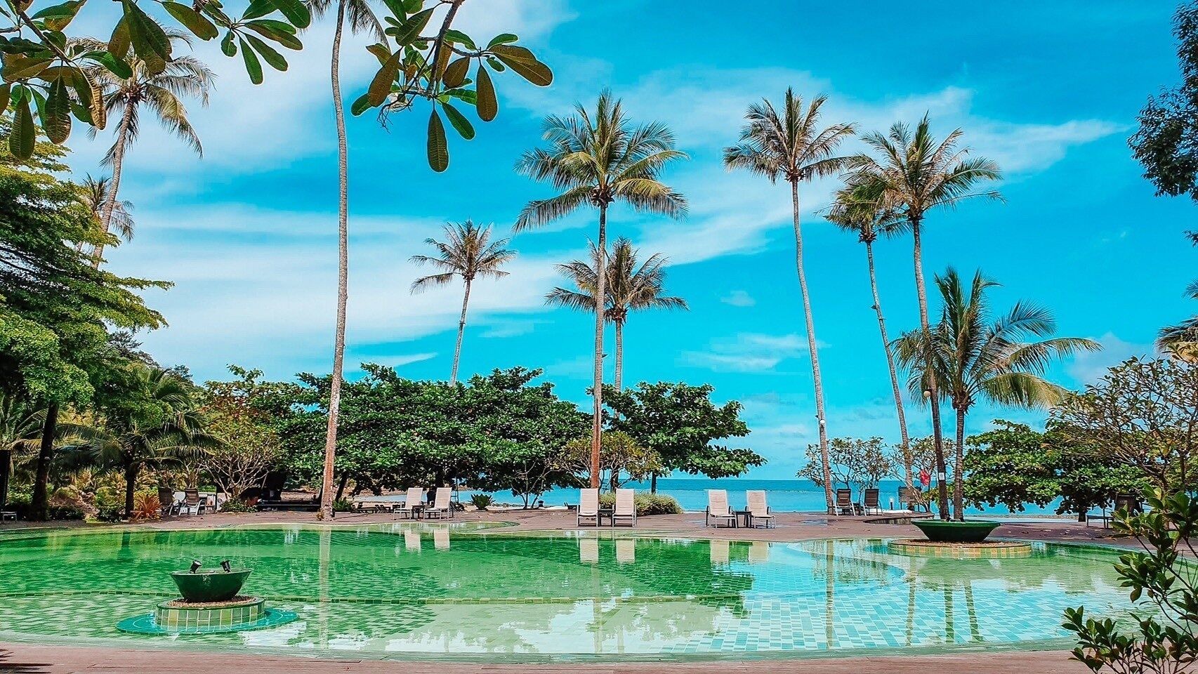 A pool area with lounge chairs and palm trees, surrounded by greenery and a clear sky.