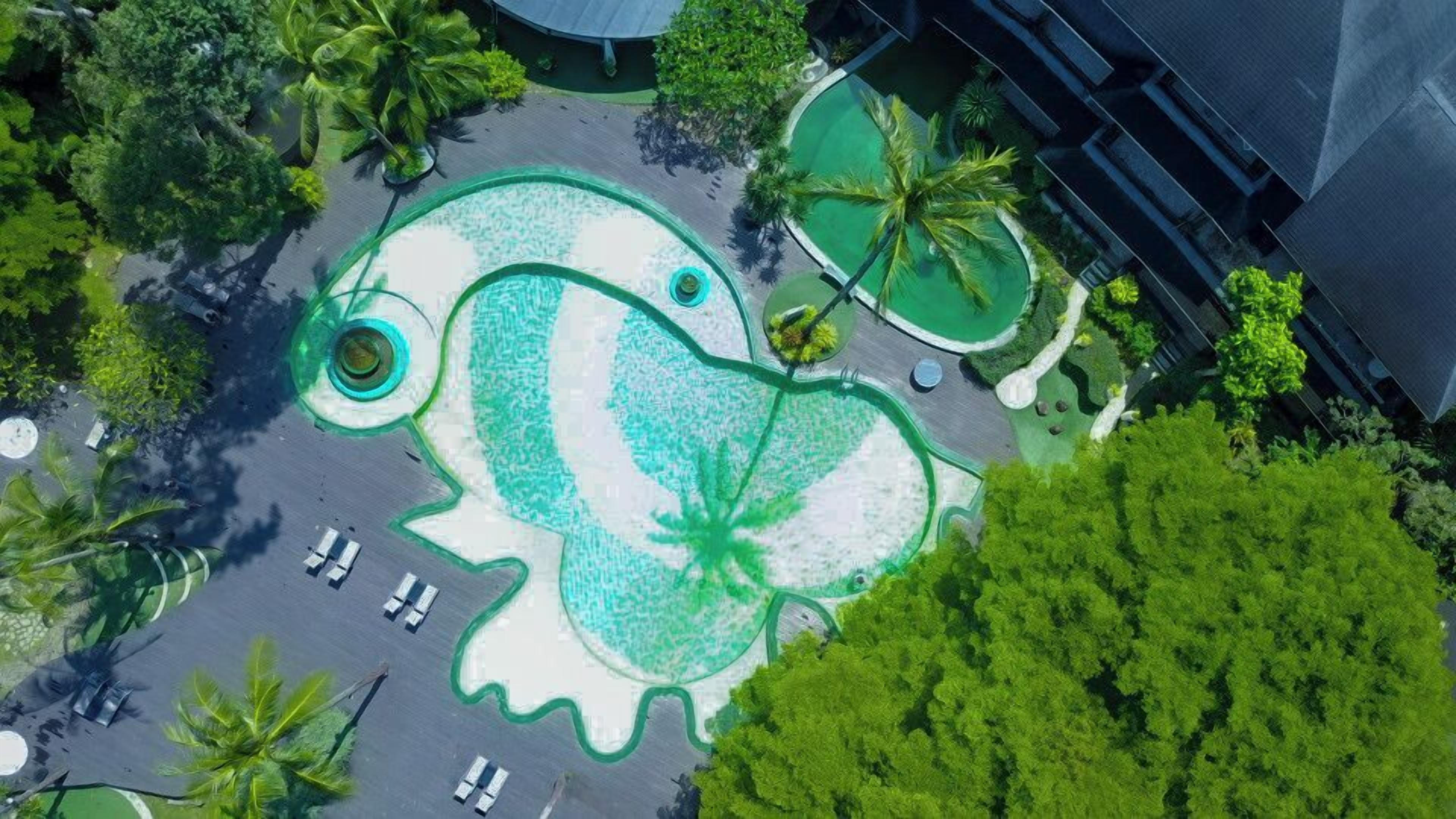 An aerial view of a resort with a large, green, frog-shaped pool surrounded by palm trees and a parking area.