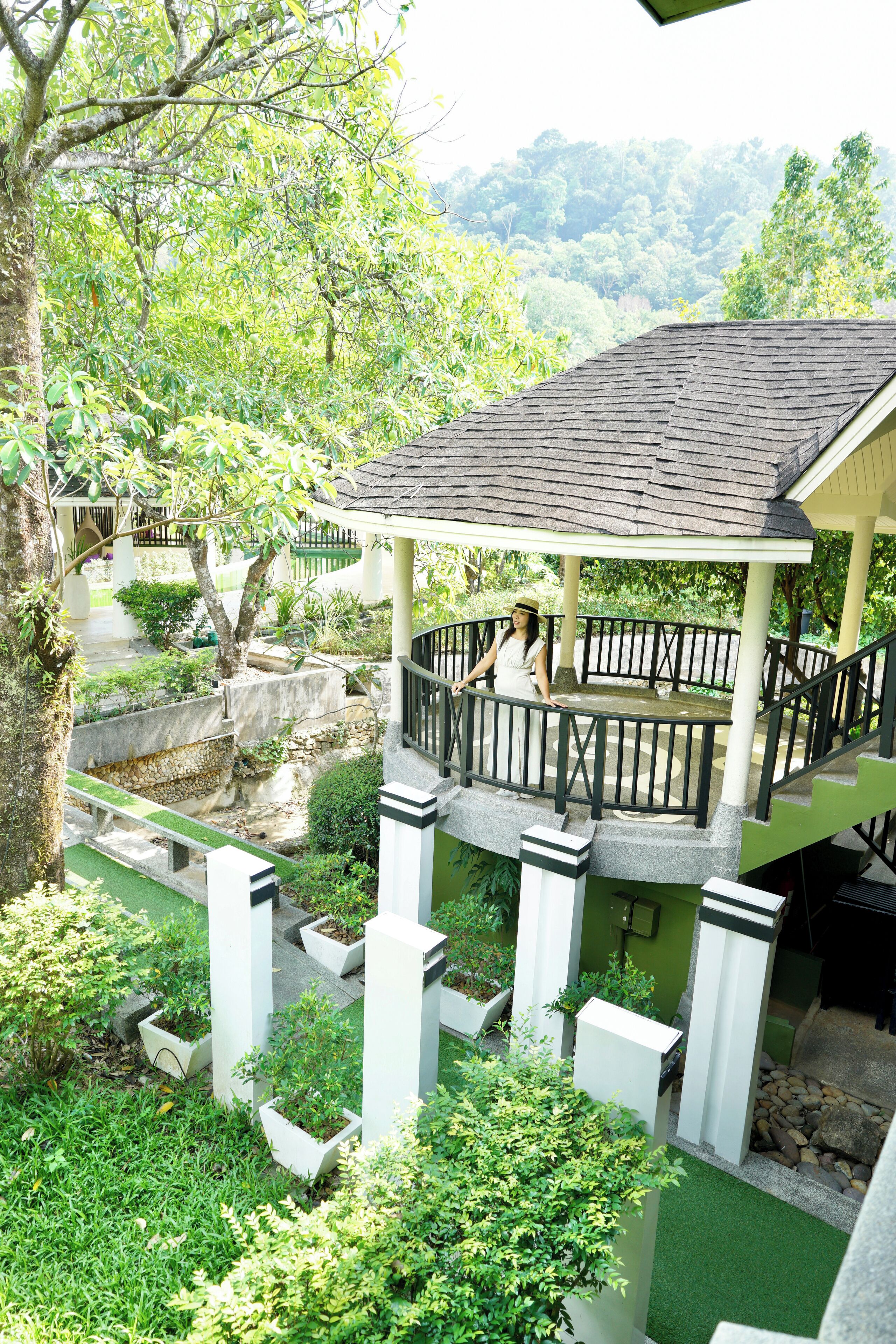 A covered outdoor area with a wooden deck, surrounded by lush greenery and a stone wall.