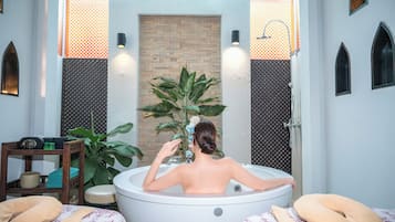Un cuarto de spa con una bañera, un estante de madera con artículos de aseo, y una planta grande en maceta.