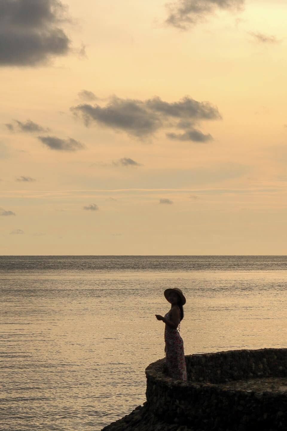 A person standing on a stone structure by the sea during sunset.