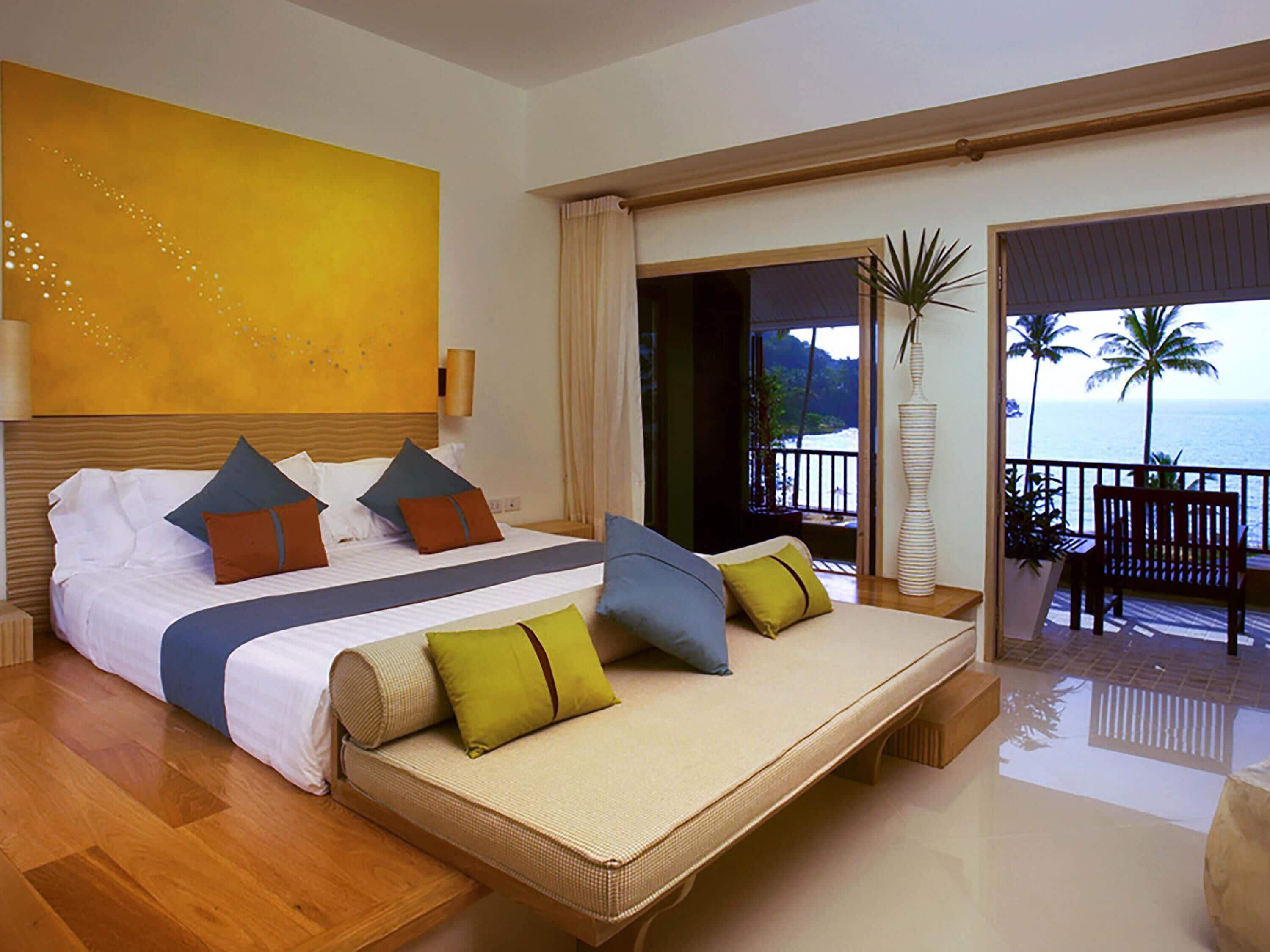 A hotel room with a bed, a balcony with a view of the ocean, and a large yellow wall.